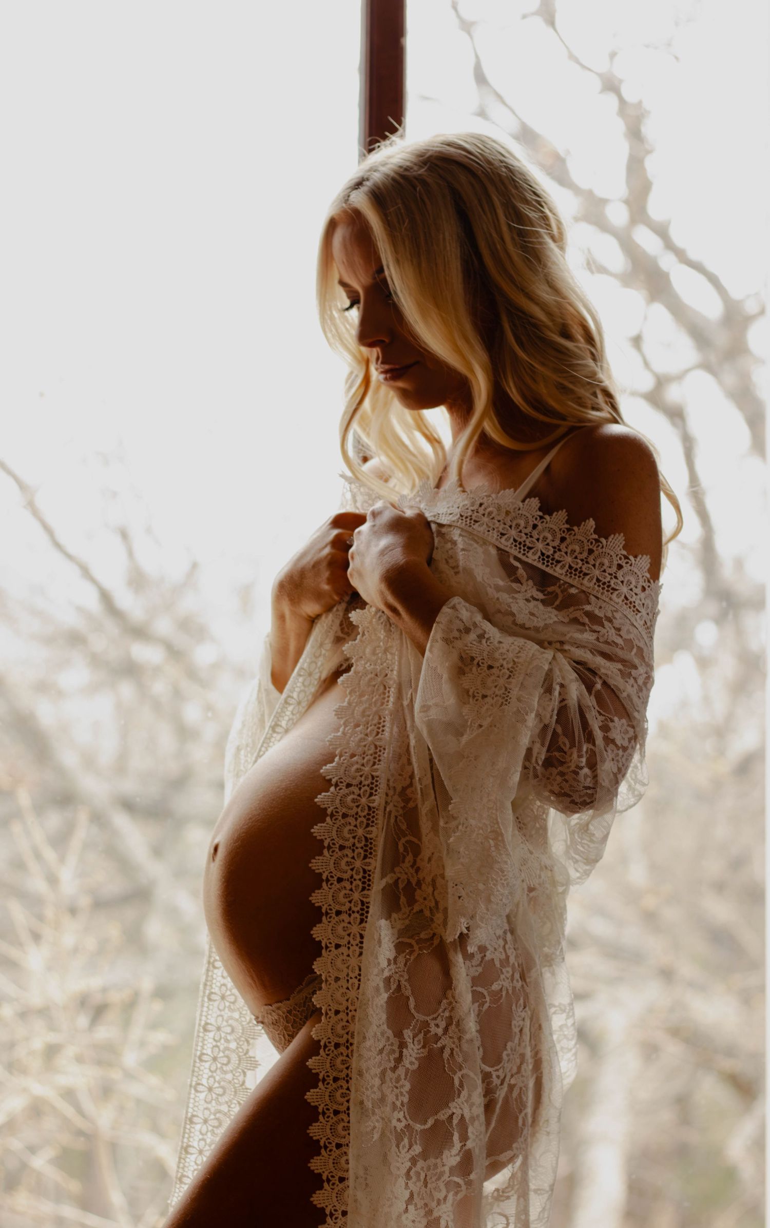 Silhouette of expectant mother in lace dress by window in soft lighting.
