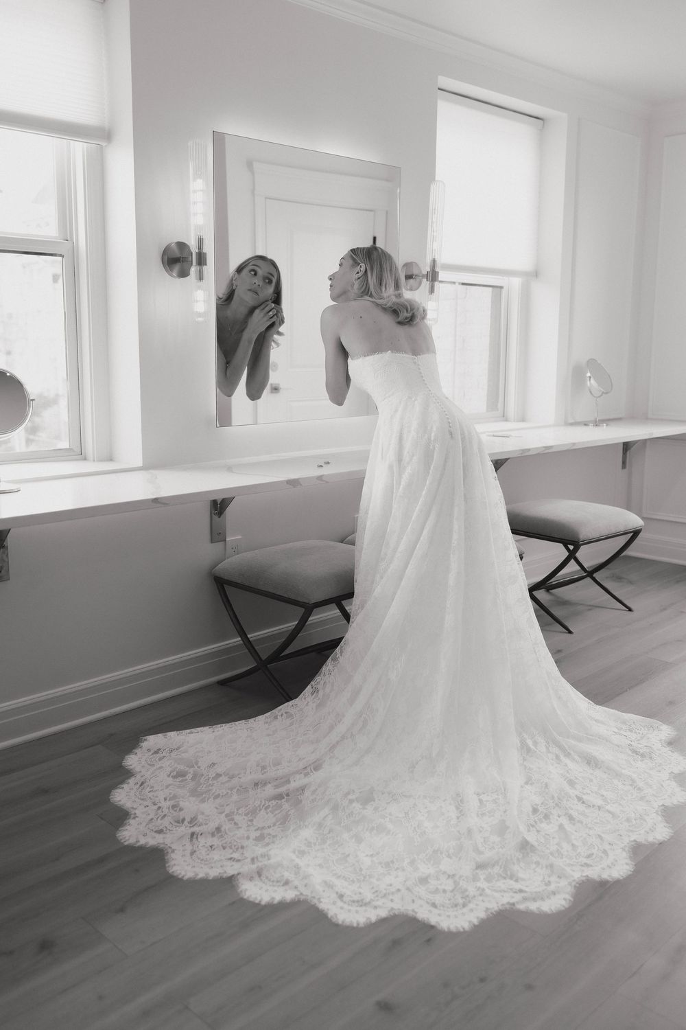 A person in an elegant white lace wedding gown with a long train looks in a mirror in a bright room with hardwood floors.