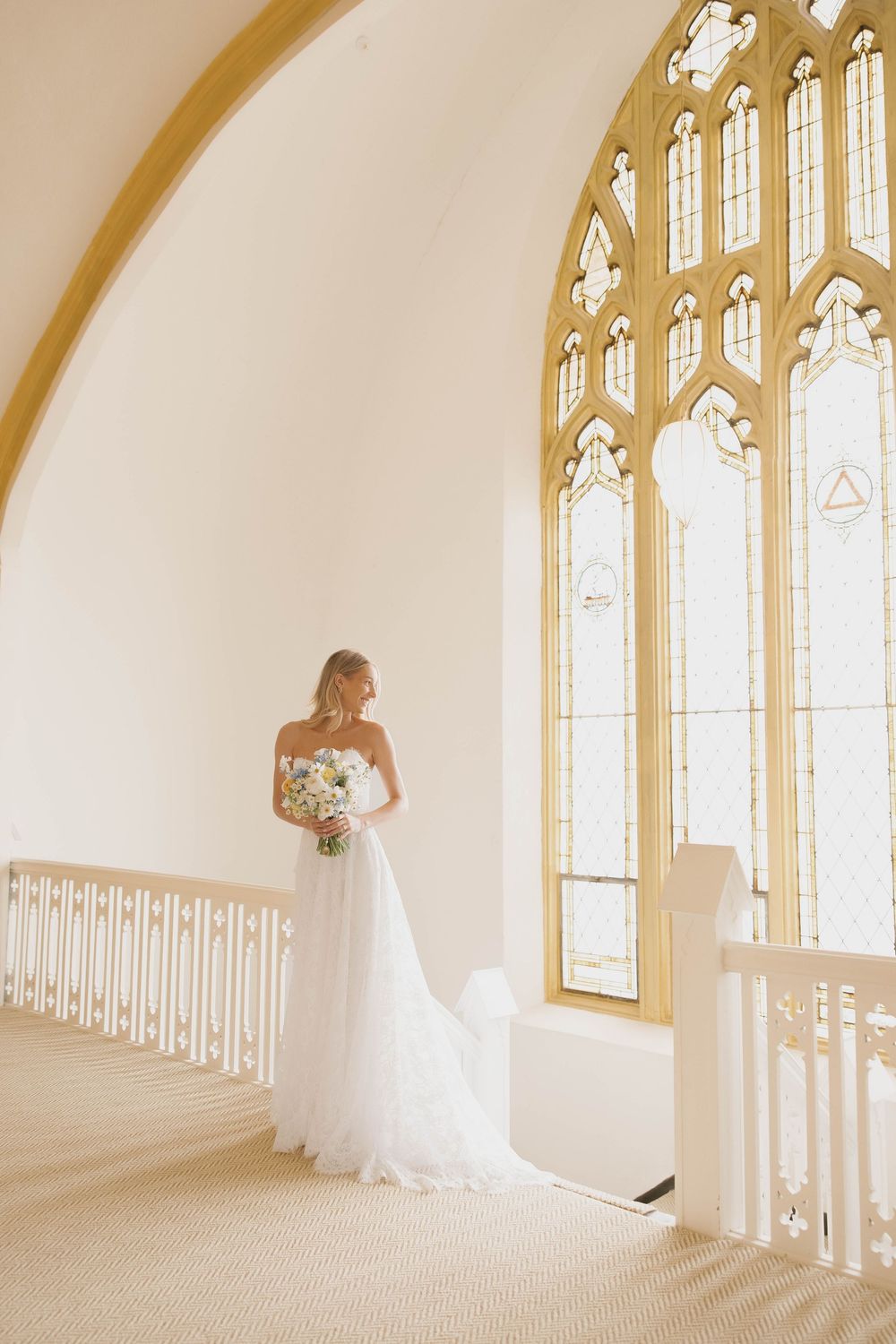 Elegant bride in white wedding dress standing alone in sunlit church sanctuary with arched windows at the Melrose Abbey in Kansas City.