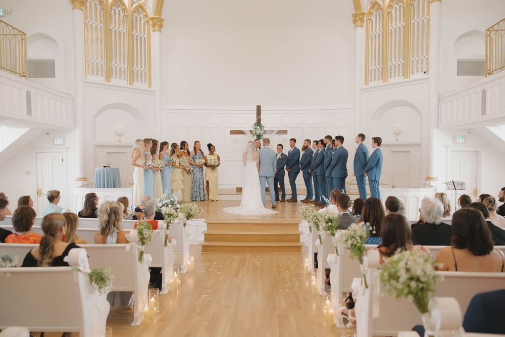 Wedding ceremony taking place in bright white church with bridal party on altar stage and guests seated in wooden pews in downtown KC.
