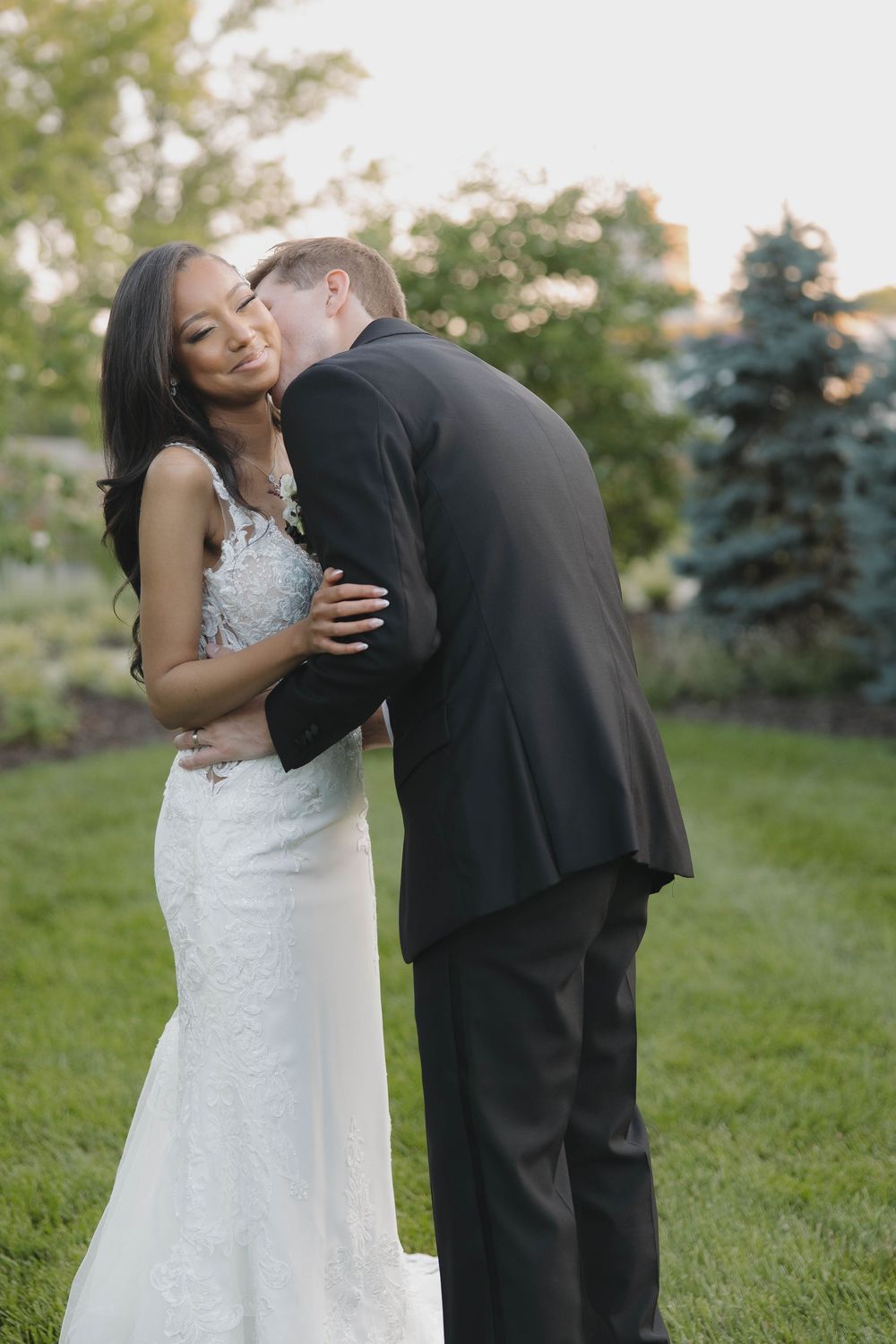 A couple shares a sweet moment on a grassy lawn with evergreen trees in the background during their wedding photoshoot.