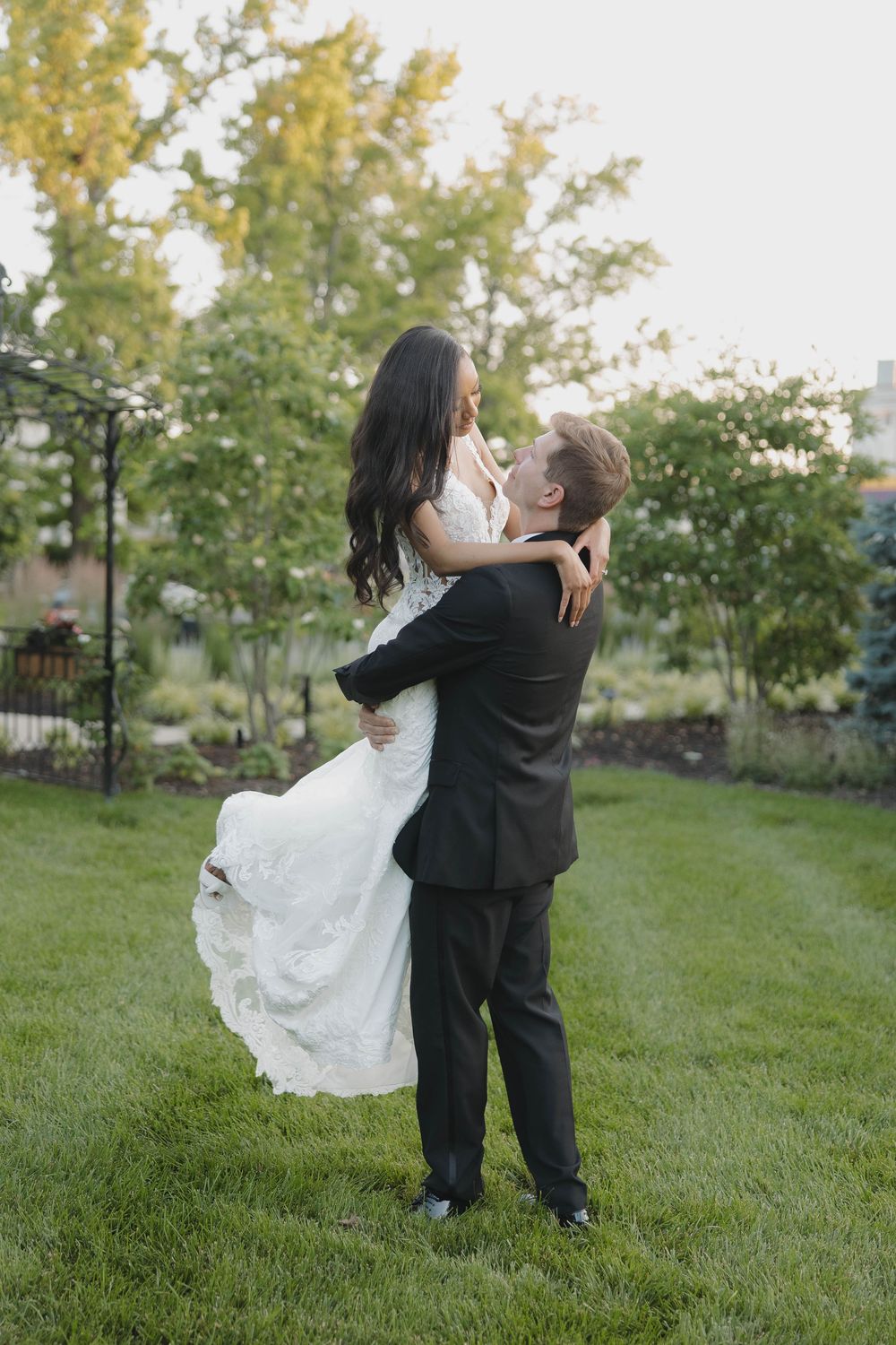 A couple in formal wedding attire embrace and dance together on a grassy lawn with trees in the background.