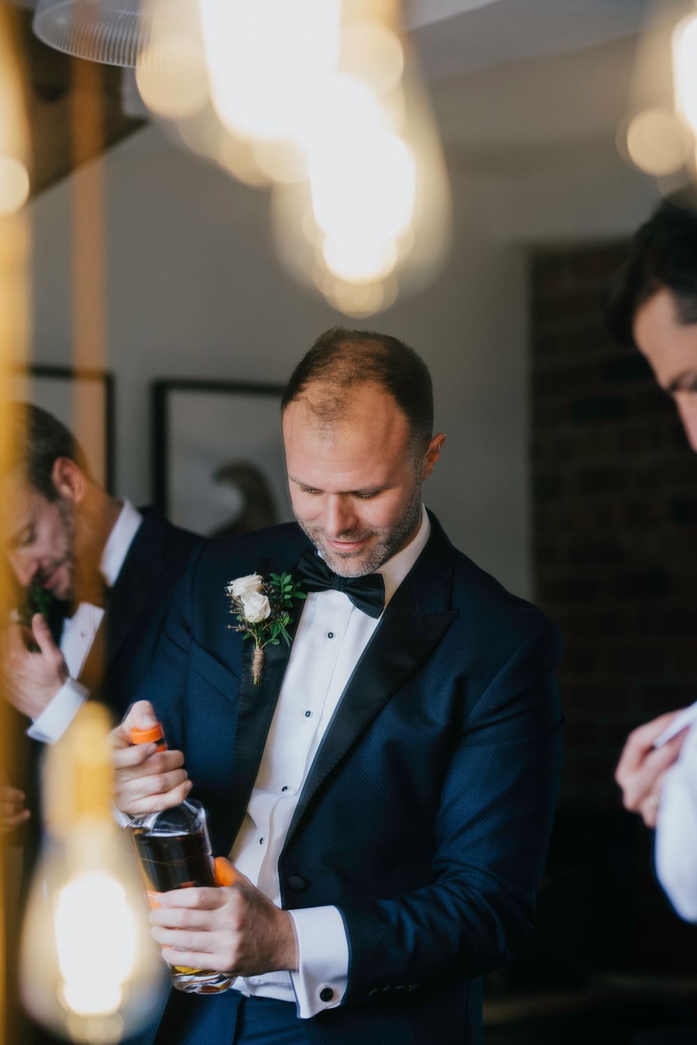 A person in a navy tuxedo and bowtie opens a bottle during an elegant event with bokeh lights in the background.