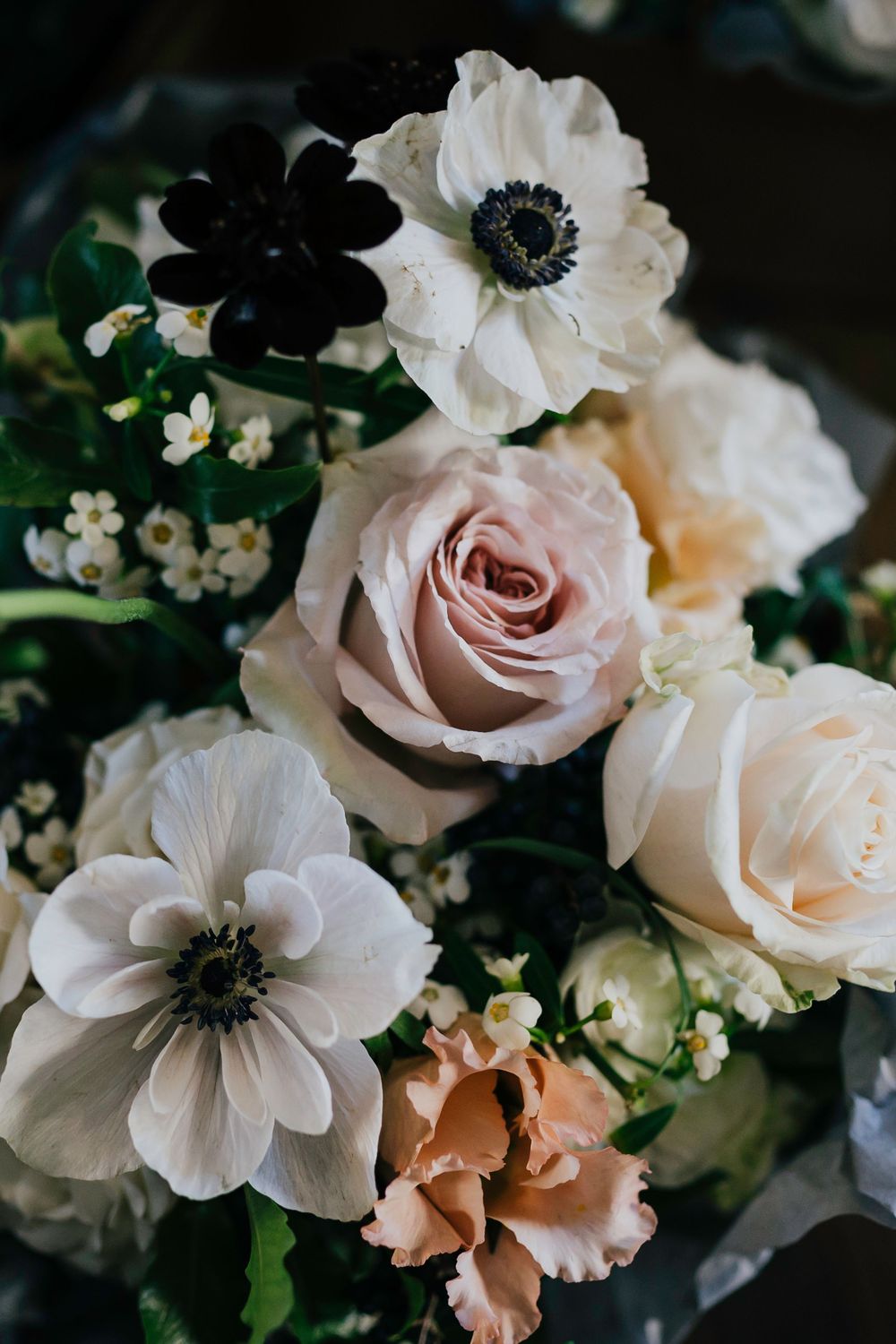 Close-up of a romantic floral arrangement with white anemones, blush roses, and coral carnations against a dark background.
