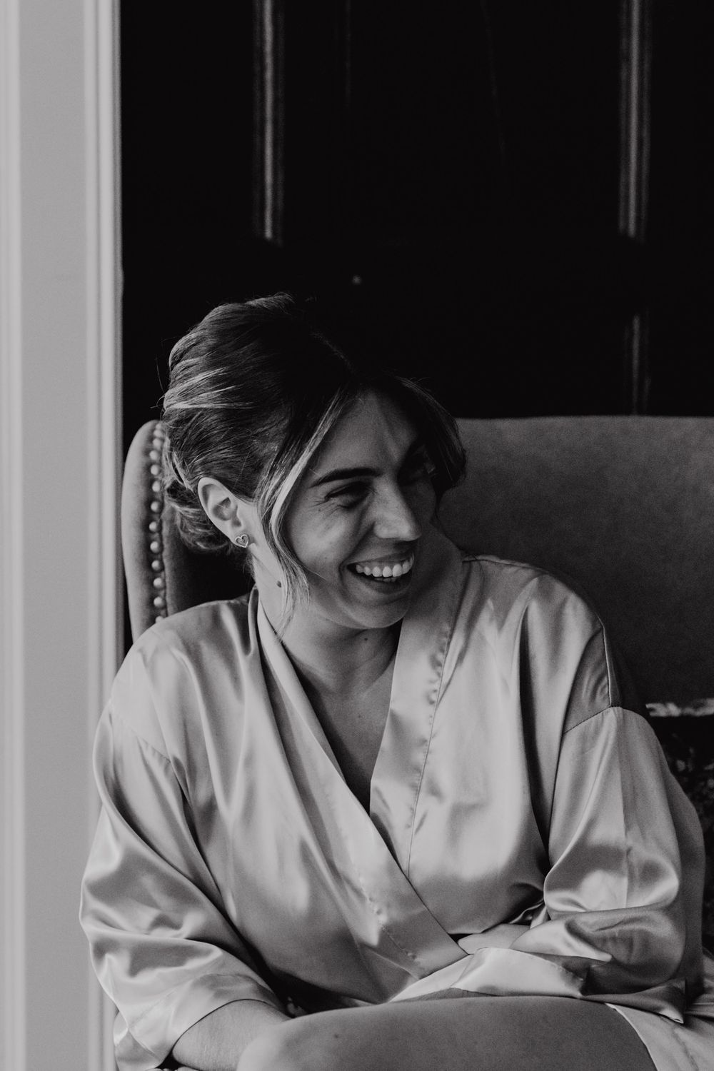Black and white photo of someone in a white silk robe sitting in a chair by a window, smiling warmly.