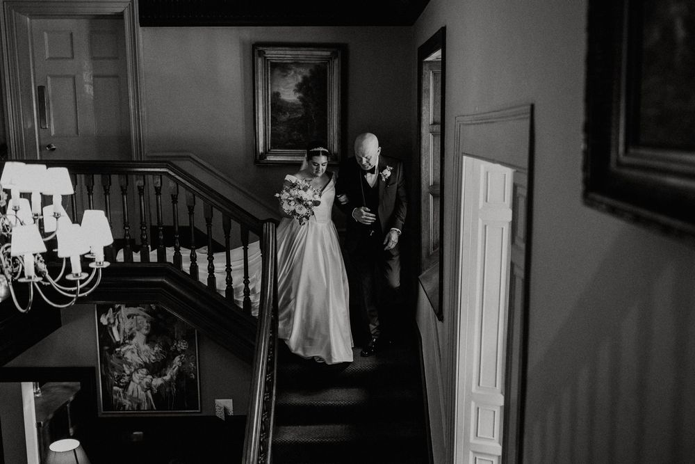 A black and white photo shows a couple descending an elegant curved staircase in a historic house with a chandelier.