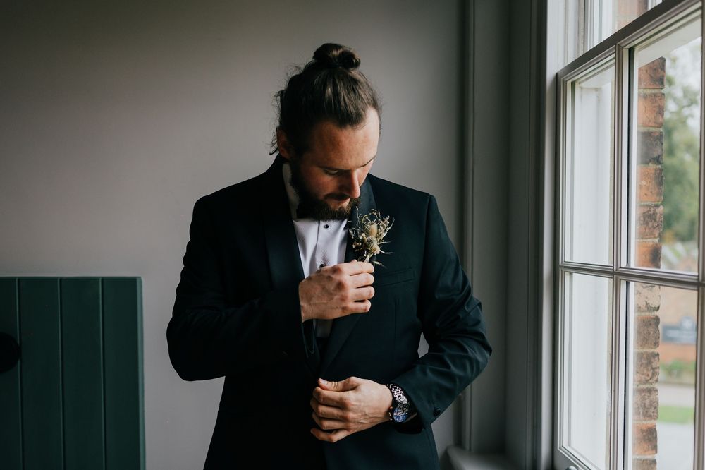 A person in a dark suit adjusts their boutonniere while standing by a window in moody lighting.