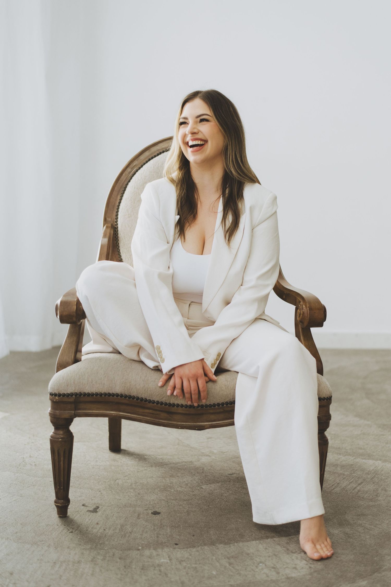 A woman in an all-white outfit laughs while sitting in an antique chair against a minimalist background.