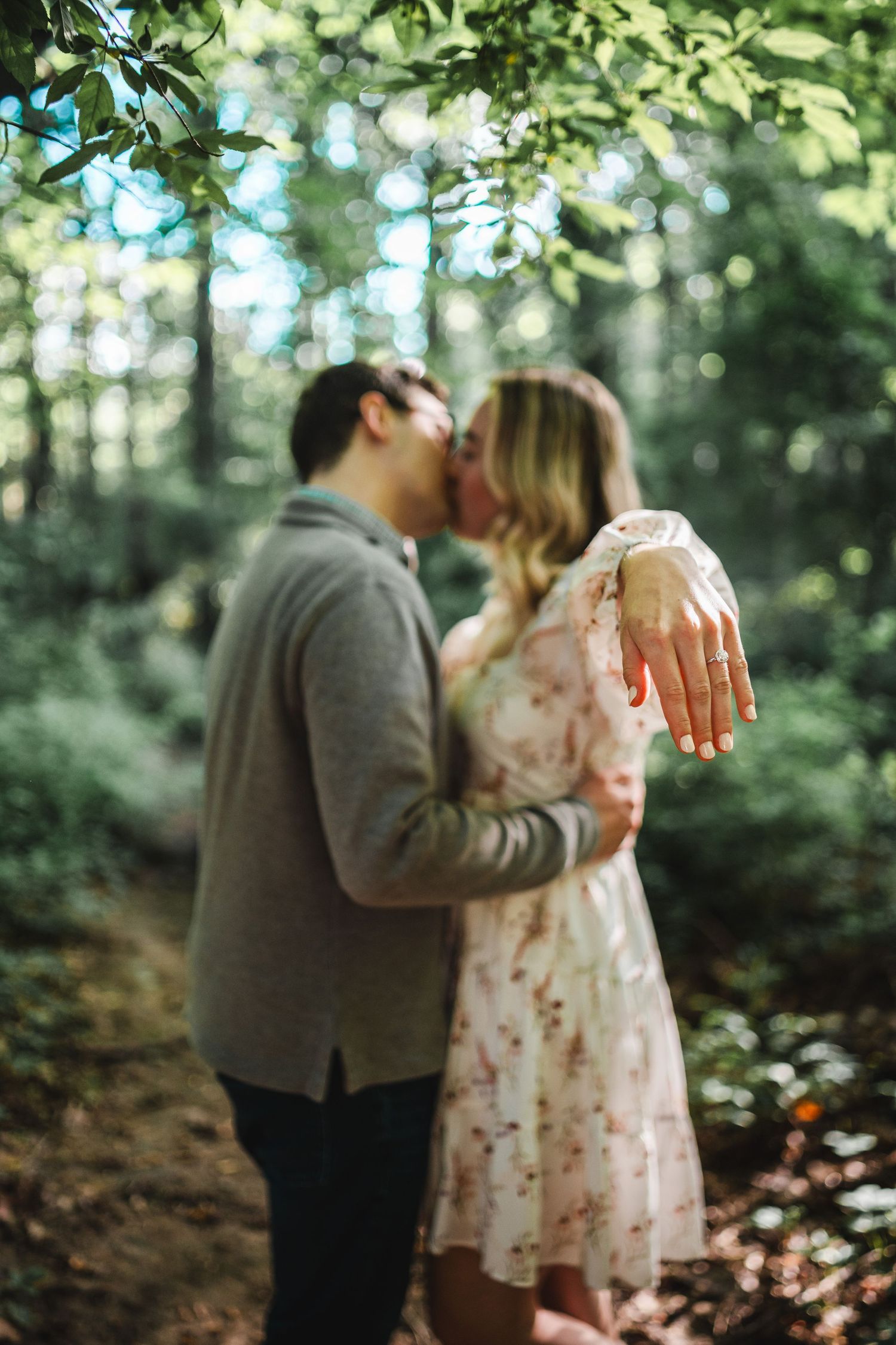 A couple shares a romantic moment in a forest while showcasing an engagement ring on the woman's hand in focus.