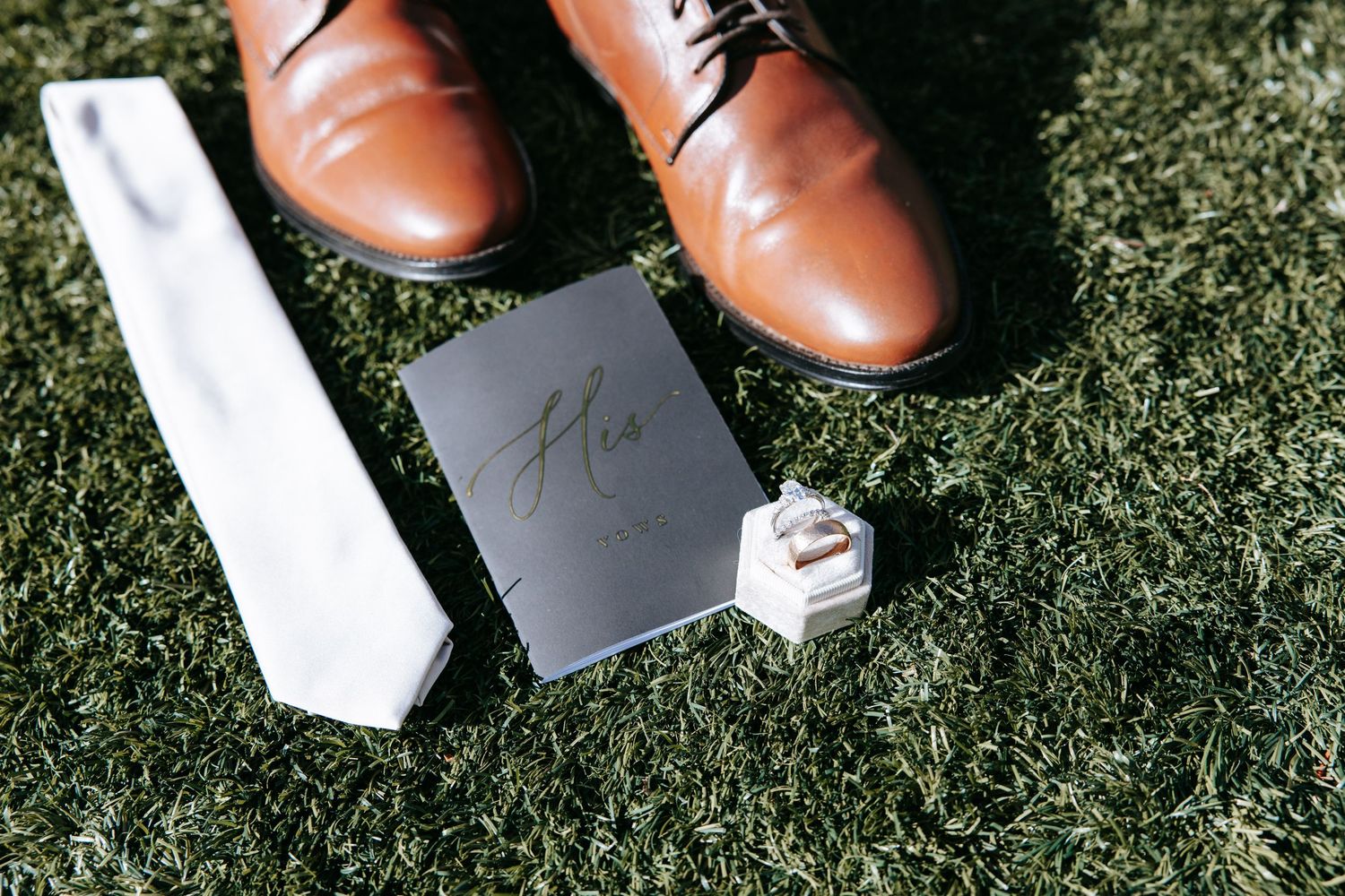 Brown leather dress shoes are placed on green grass next to a white surface and gray card.