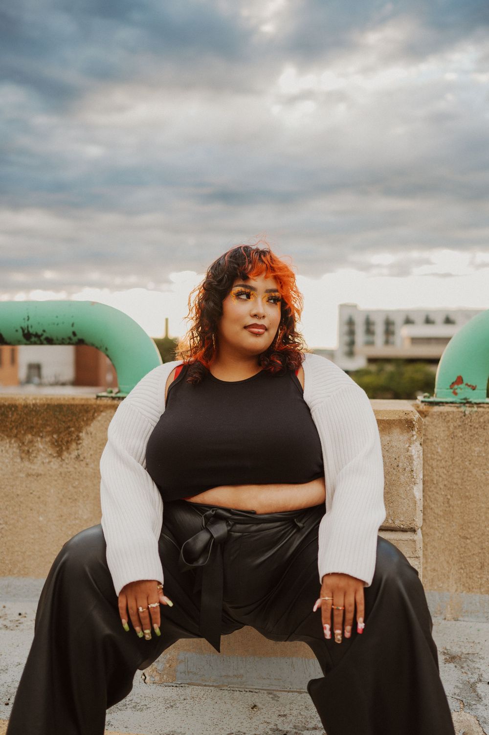 A curly-haired model in black and white outfit poses on a rooftop during golden hour with cloudy skies.