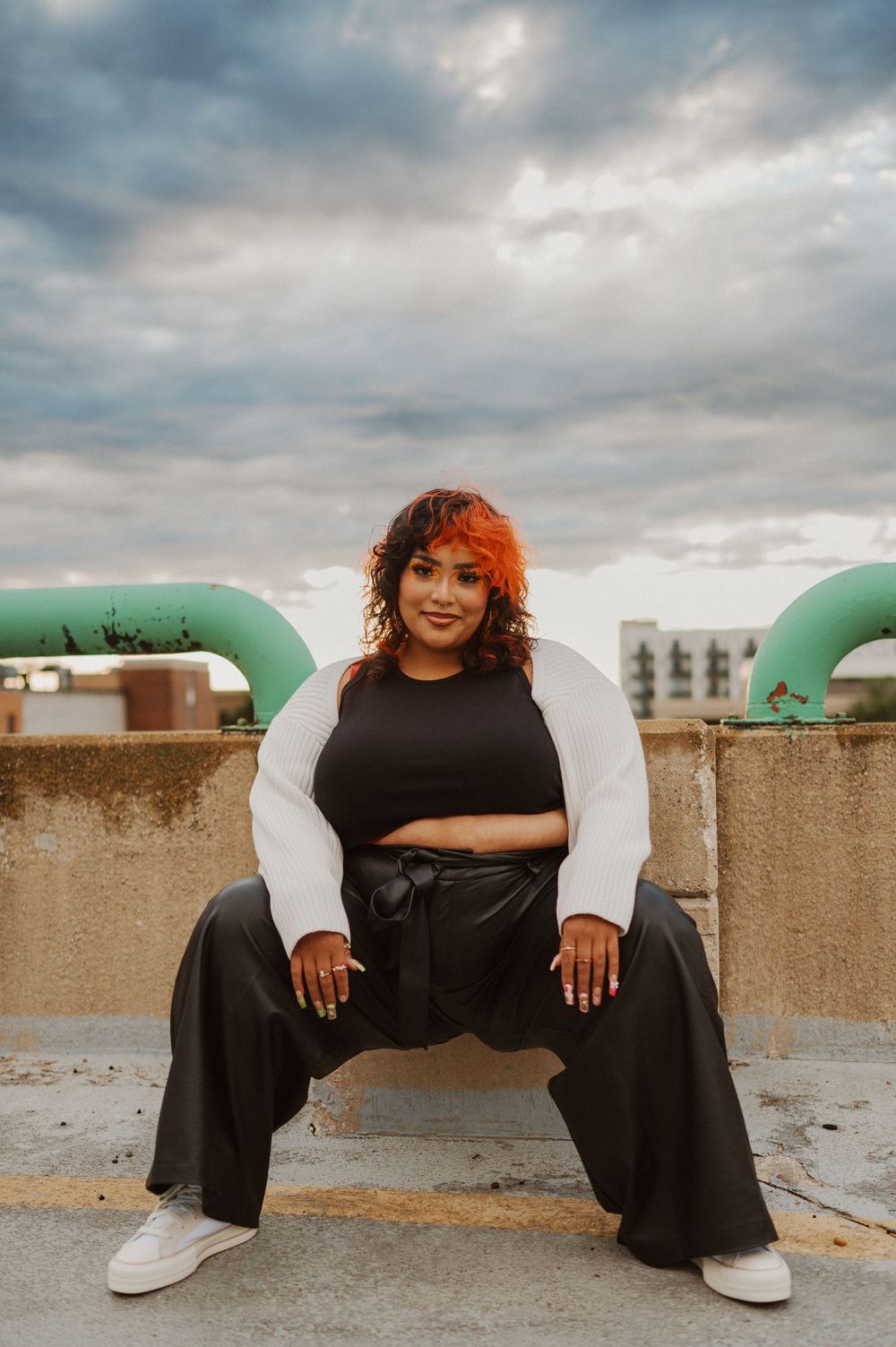 A person in black attire poses crouched between large green pipes on a rooftop under dramatic cloudy skies.