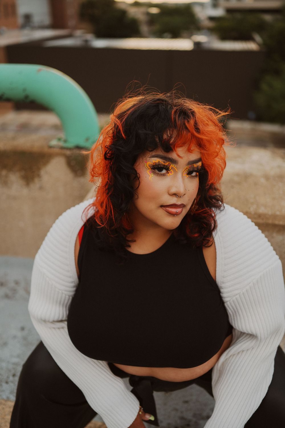 Artistic portrait of someone with vibrant orange hair wearing a black crop top and white long sleeve shrug outdoors.