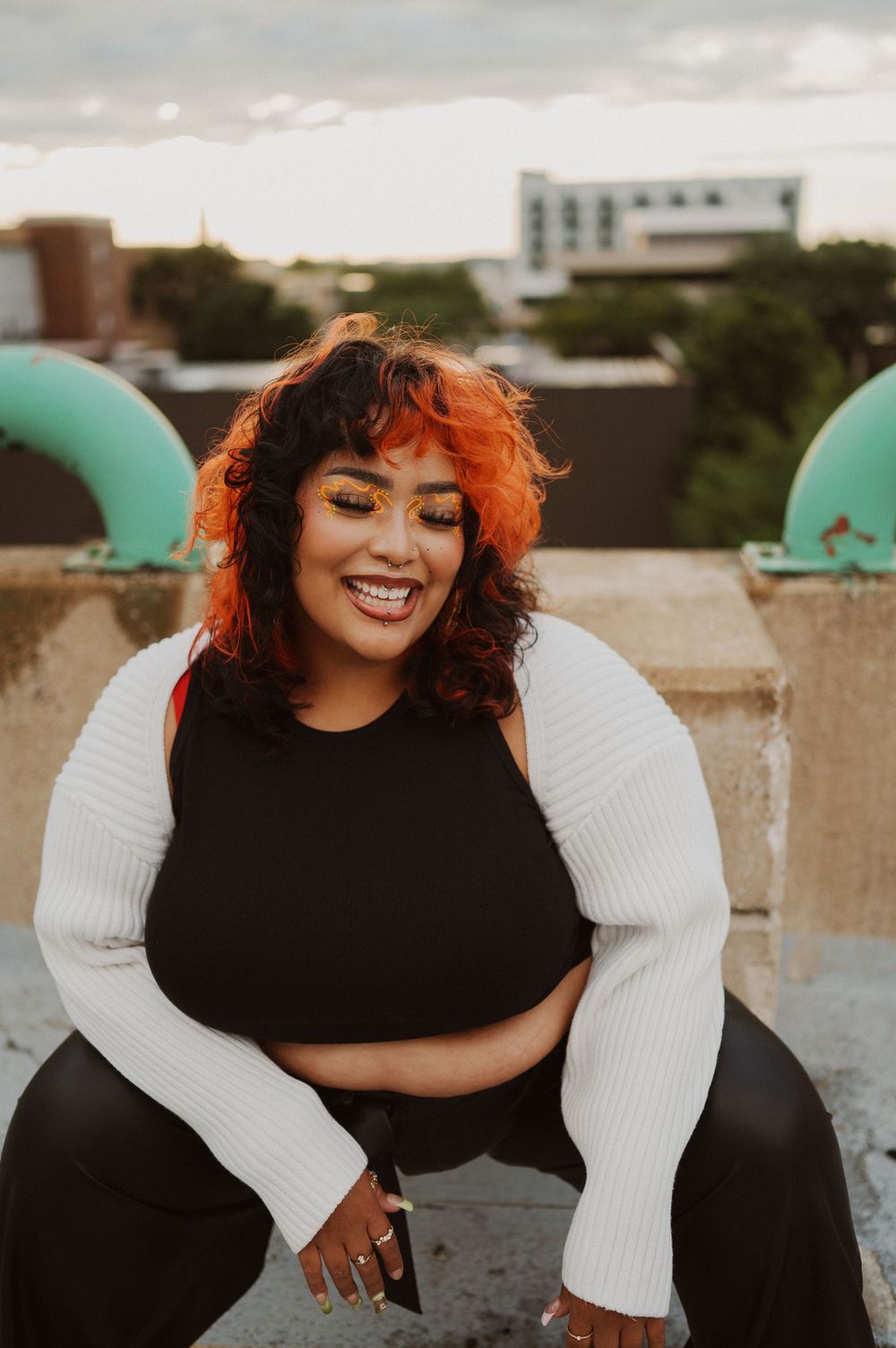 A person with vibrant orange hair wearing a black crop top and white sleeves sits smiling on a rooftop at sunset.