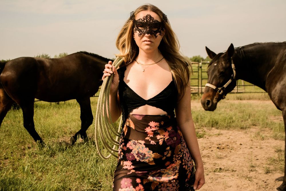 A person in a black mask and floral dress stands with horses on a dirt path in a sunny field.
