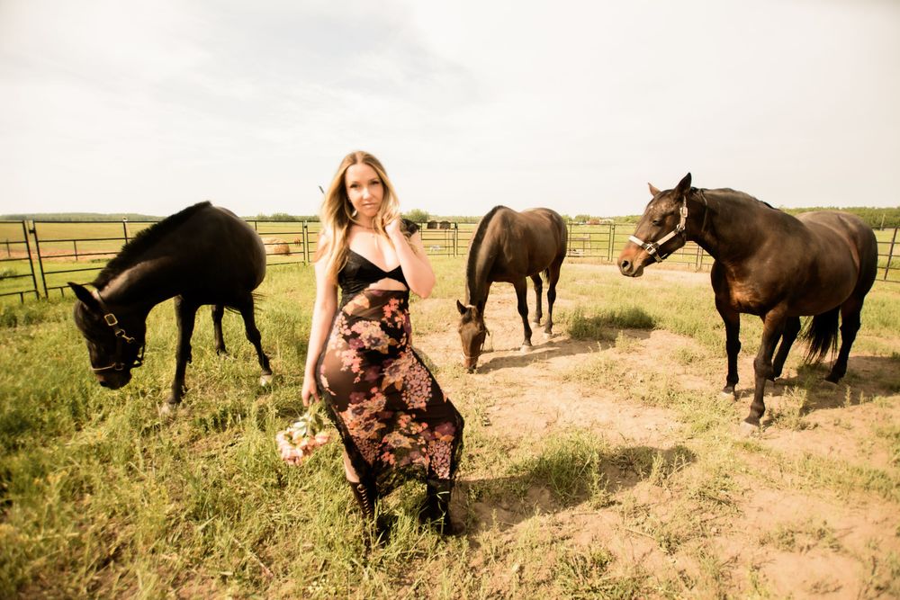Multiple horses graze in a fenced pasture while someone in a floral dress poses in the foreground.
