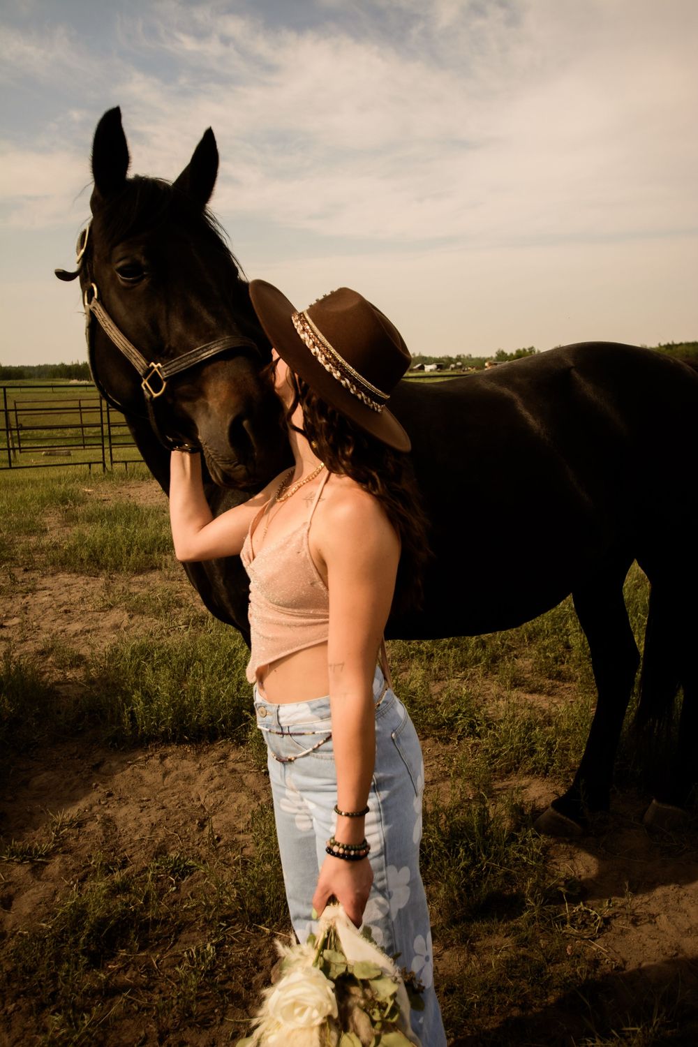 A western-styled photoshoot featuring a dark horse and a person in a cowboy hat and casual attire in a rustic field setting.