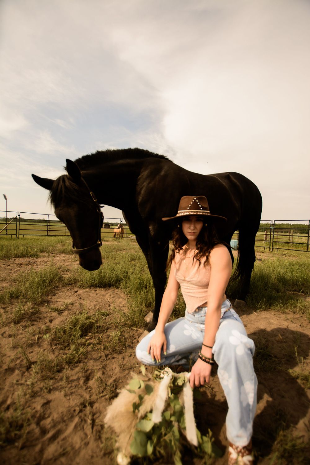 A black horse grazes next to a person wearing a cowboy hat and jeans in a rustic outdoor setting at sunset.