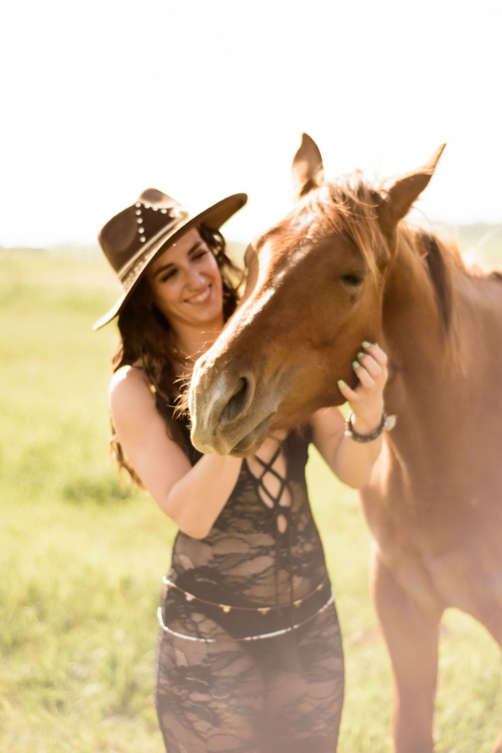 A woman in western attire lovingly embraces her brown horse in a sunlit field.
