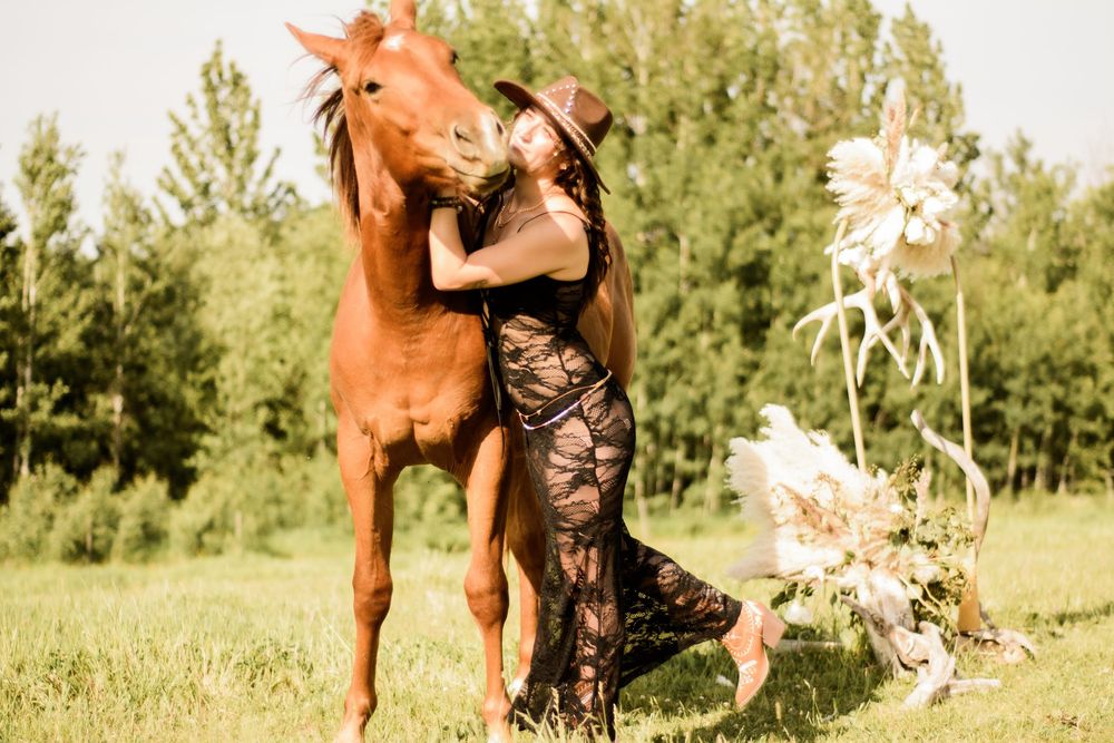 A chestnut horse and someone in a black lace outfit pose together in a sunny field with trees.