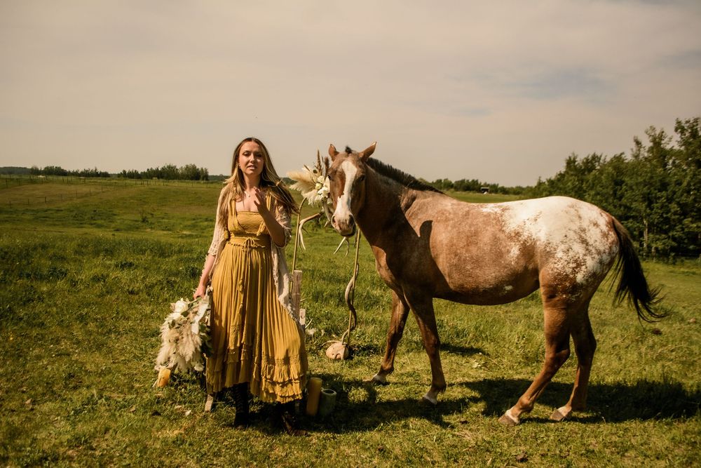A spotted horse stands beside someone wearing a flowing yellow dress in a green pasture.