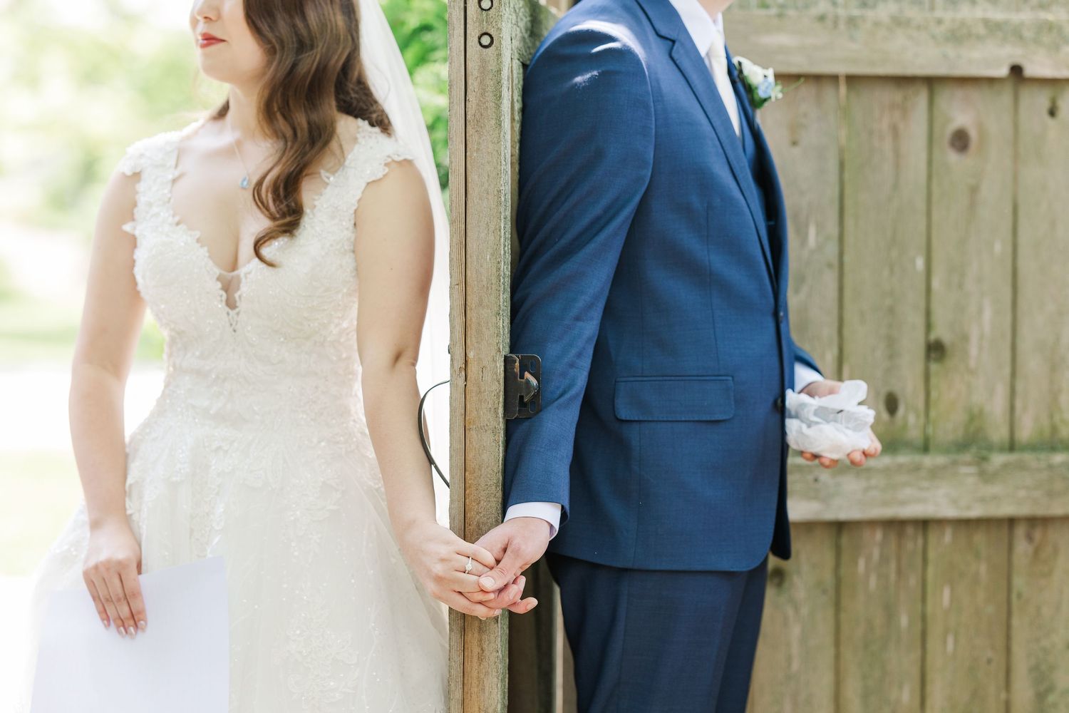 Bride and groom hold hands while standing on opposite sides of rustic wooden fence during first look photo.