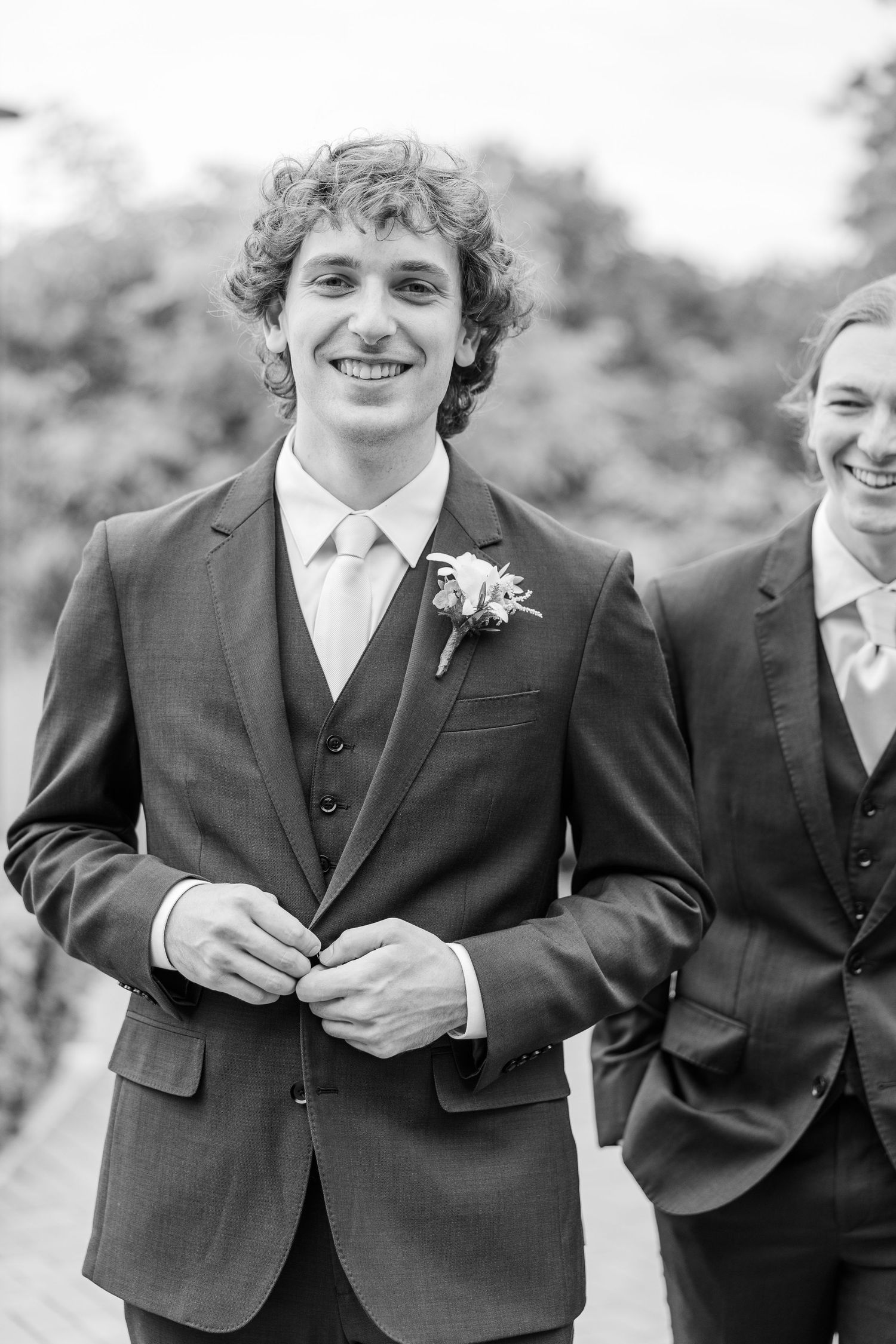 Groomsmen in matching navy blue suits and brown shoes share a lighthearted moment on a brick pathway surrounded by greenery.