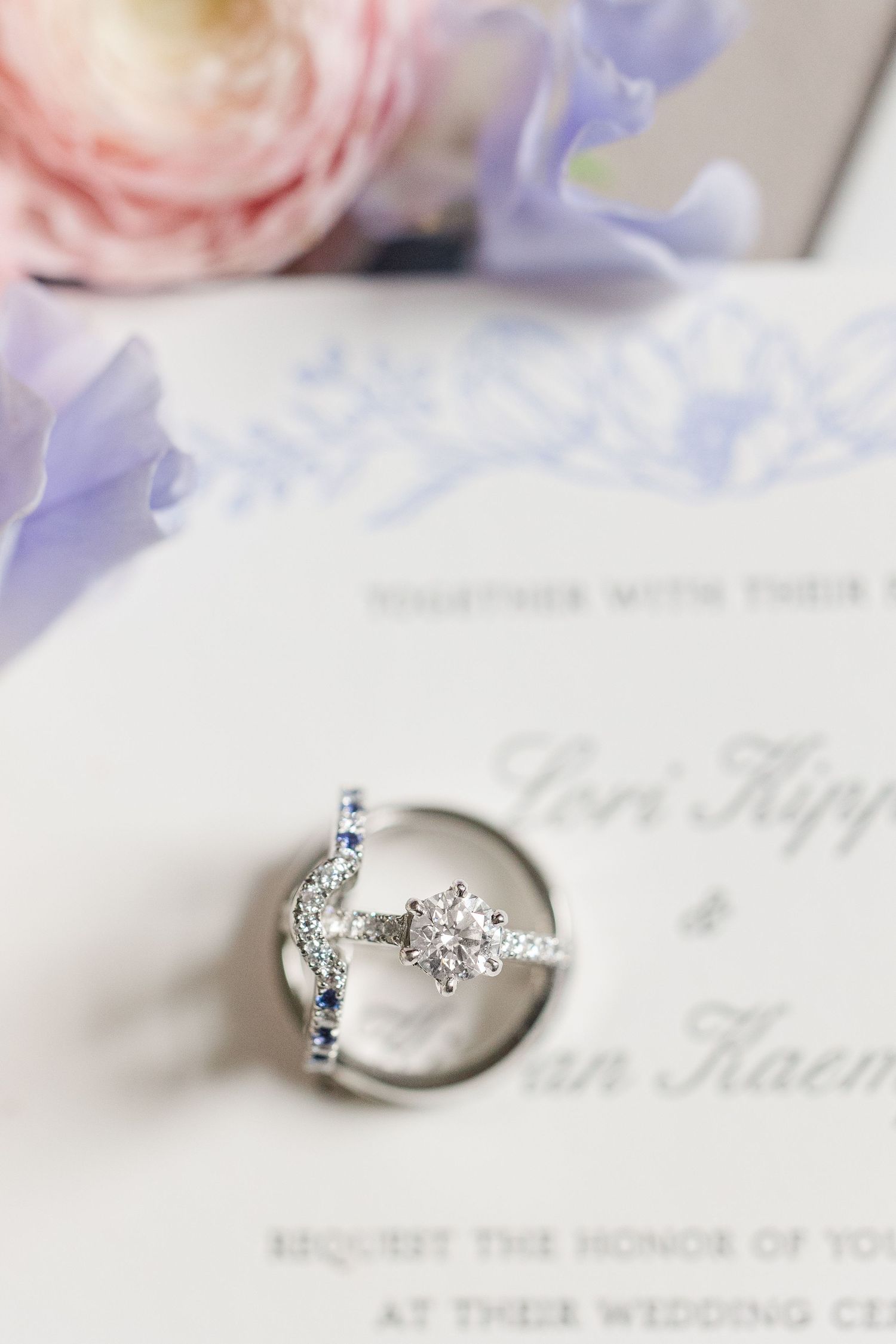 A sparkling diamond engagement ring sits on a wedding invitation with soft pink and purple flowers in the background.