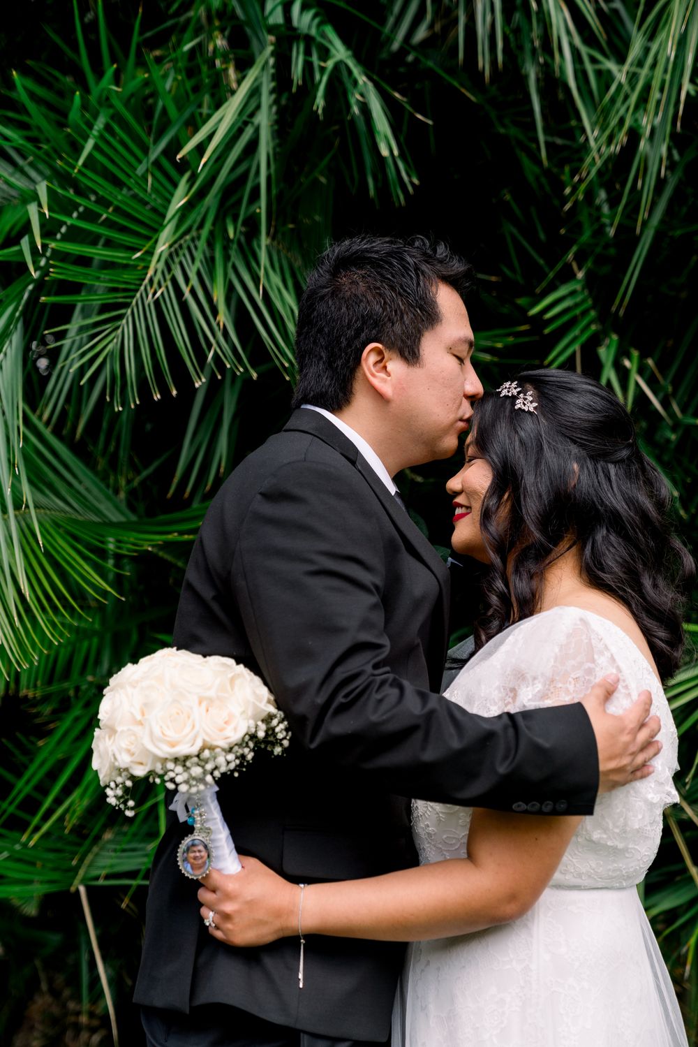 Bride and groom share a romantic embrace in front of lush tropical palm fronds during wedding photo session.