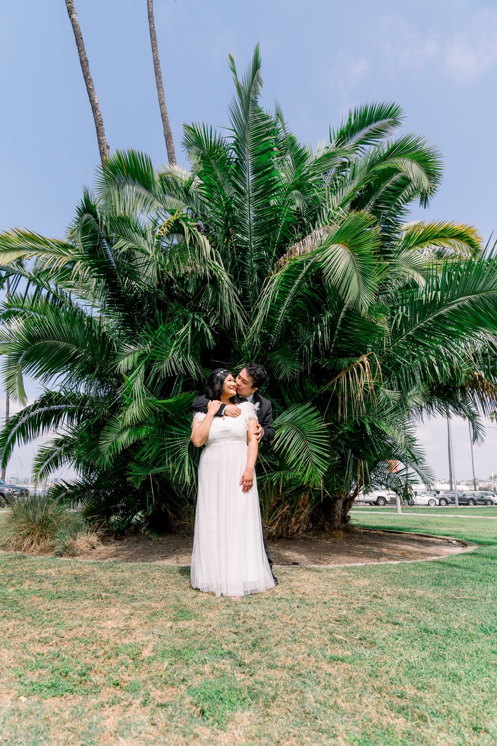 A romantic couple embraces in front of a lush palm tree on a sunny day, with the bride wearing a flowing white dress.