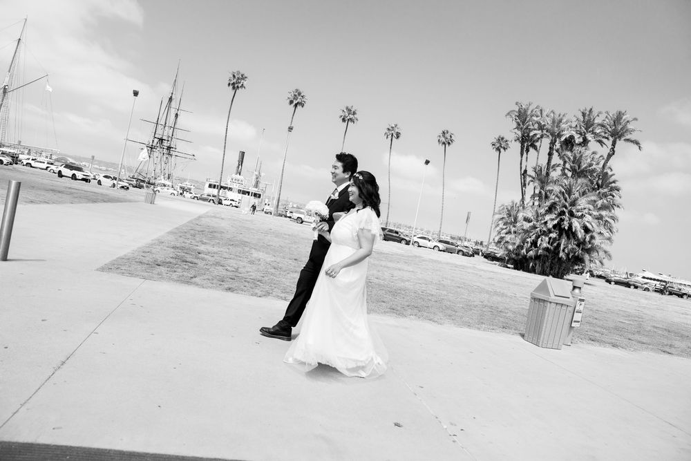 A couple in wedding attire share a romantic dance on a waterfront promenade with palm trees in the background in black and white.