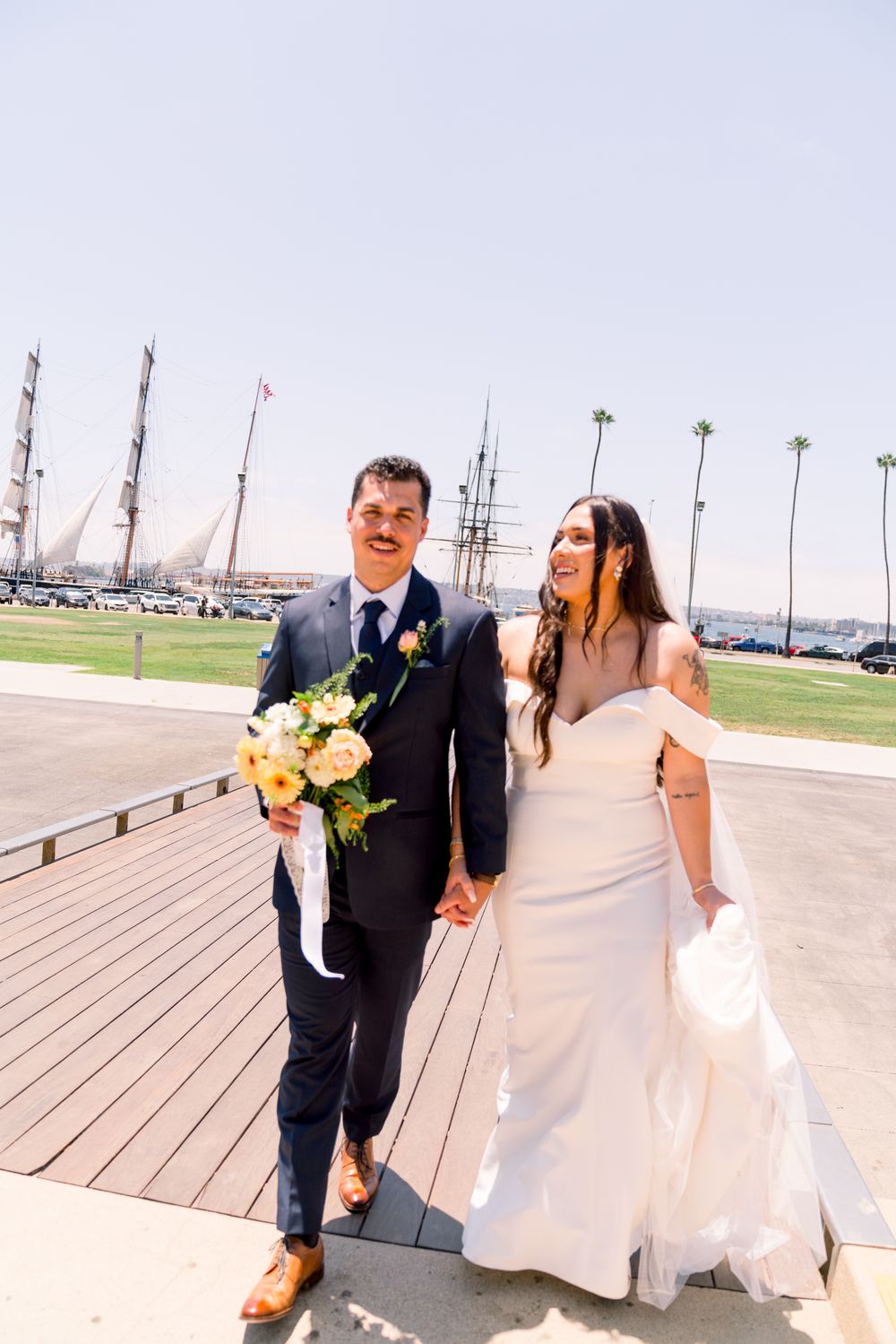 A couple in wedding attire walks along a boardwalk with palm trees and boats visible in the harbor behind them.
