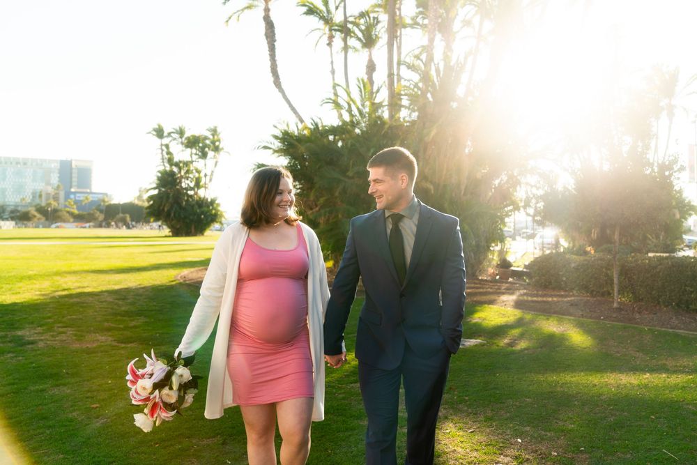 A couple walks together in a sunny park at golden hour, with the pregnant woman wearing a pink dress and white cardigan.