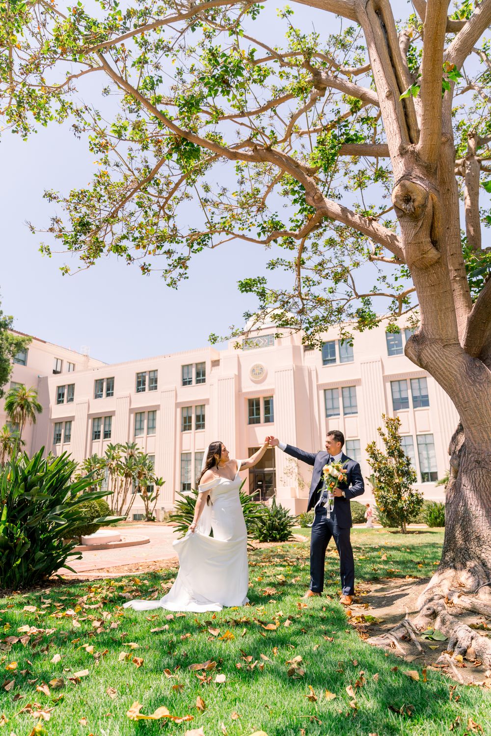 A newlywed couple takes wedding photos under large trees on a sunny green lawn outside a light pink building.