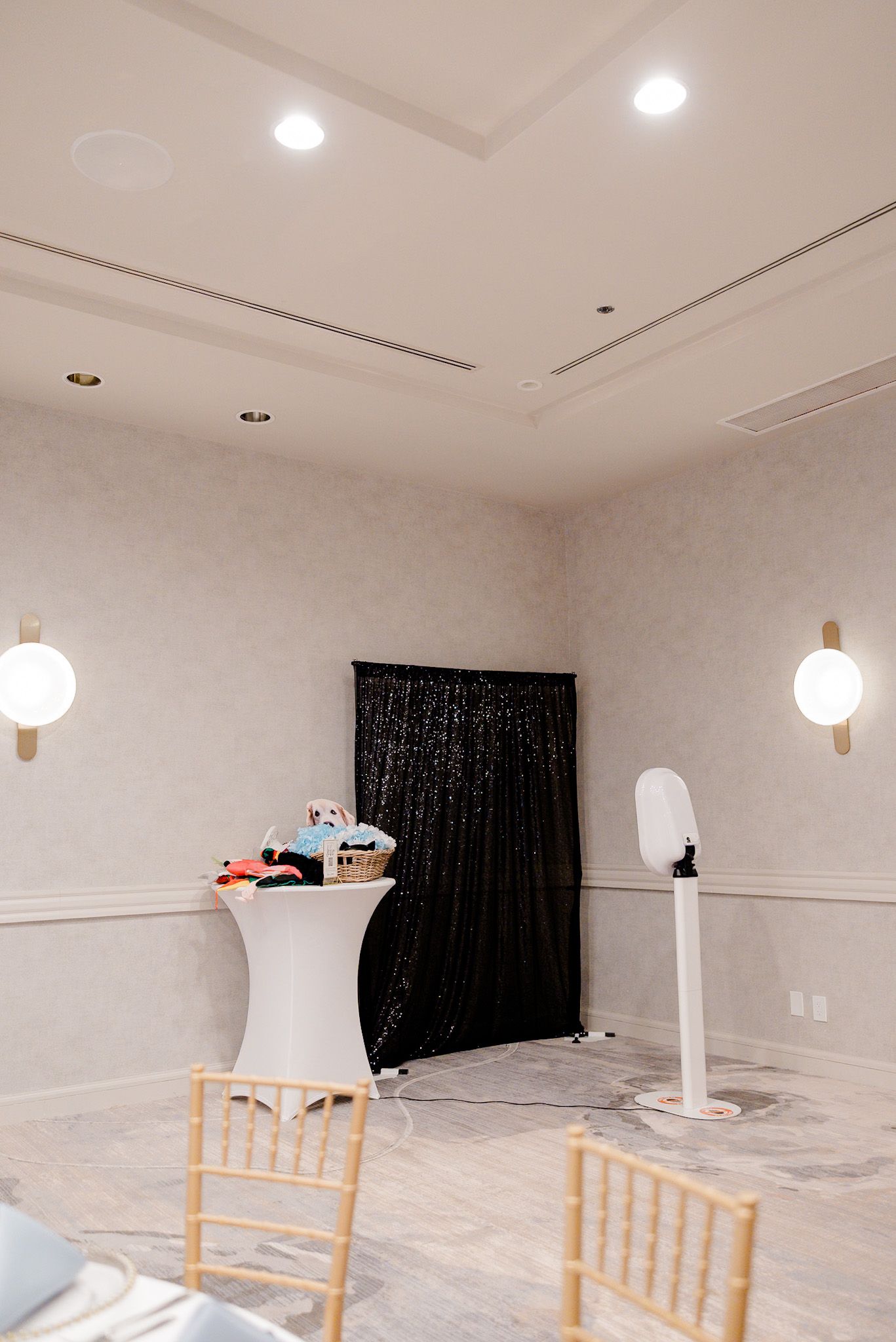 Photo booth setup with black sequin backdrop, white cocktail table and wooden chiavari chairs in an elegant event space.
