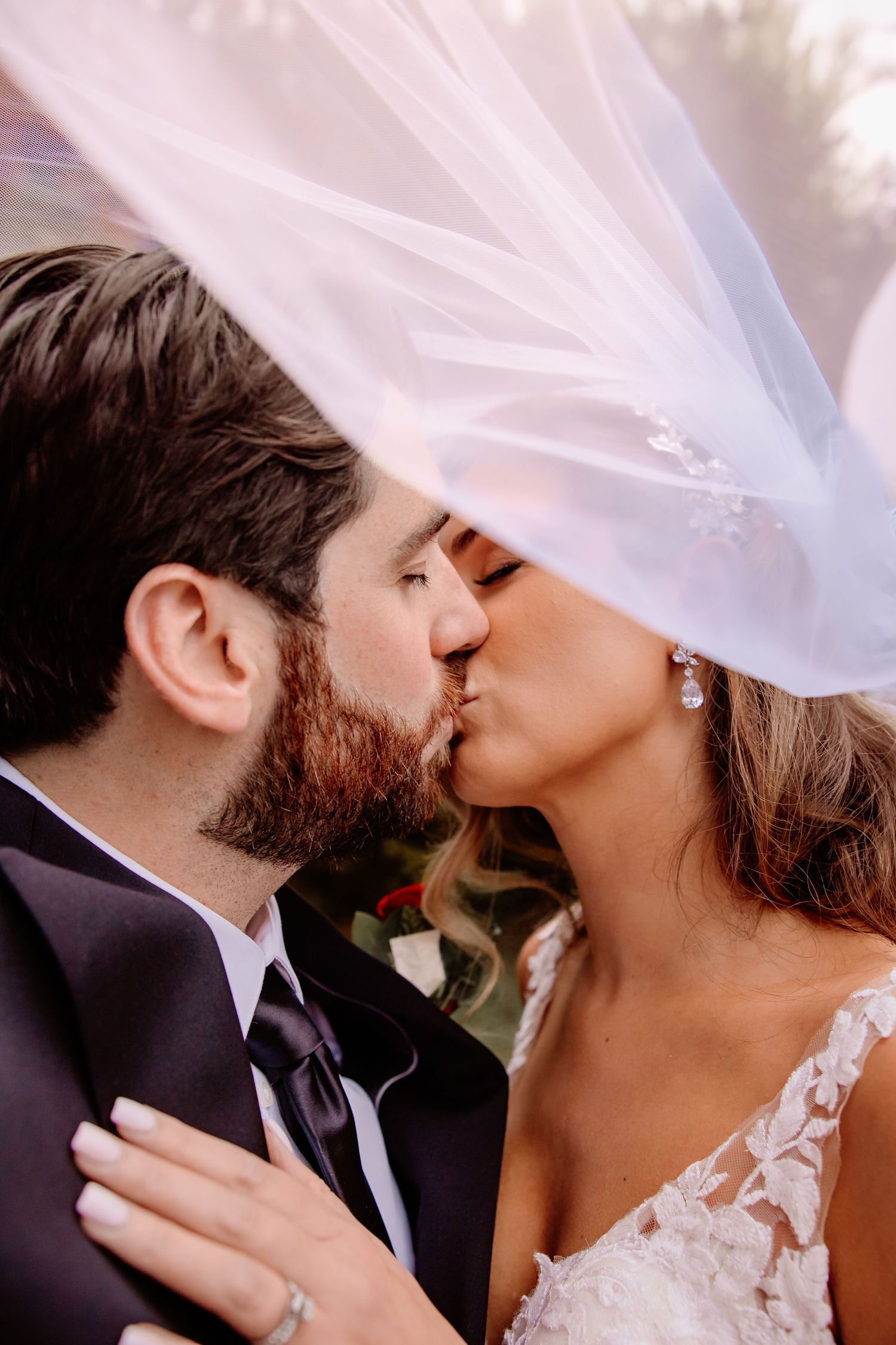 A romantic wedding moment captured under a flowing veil as a couple shares their first kiss as newlyweds.