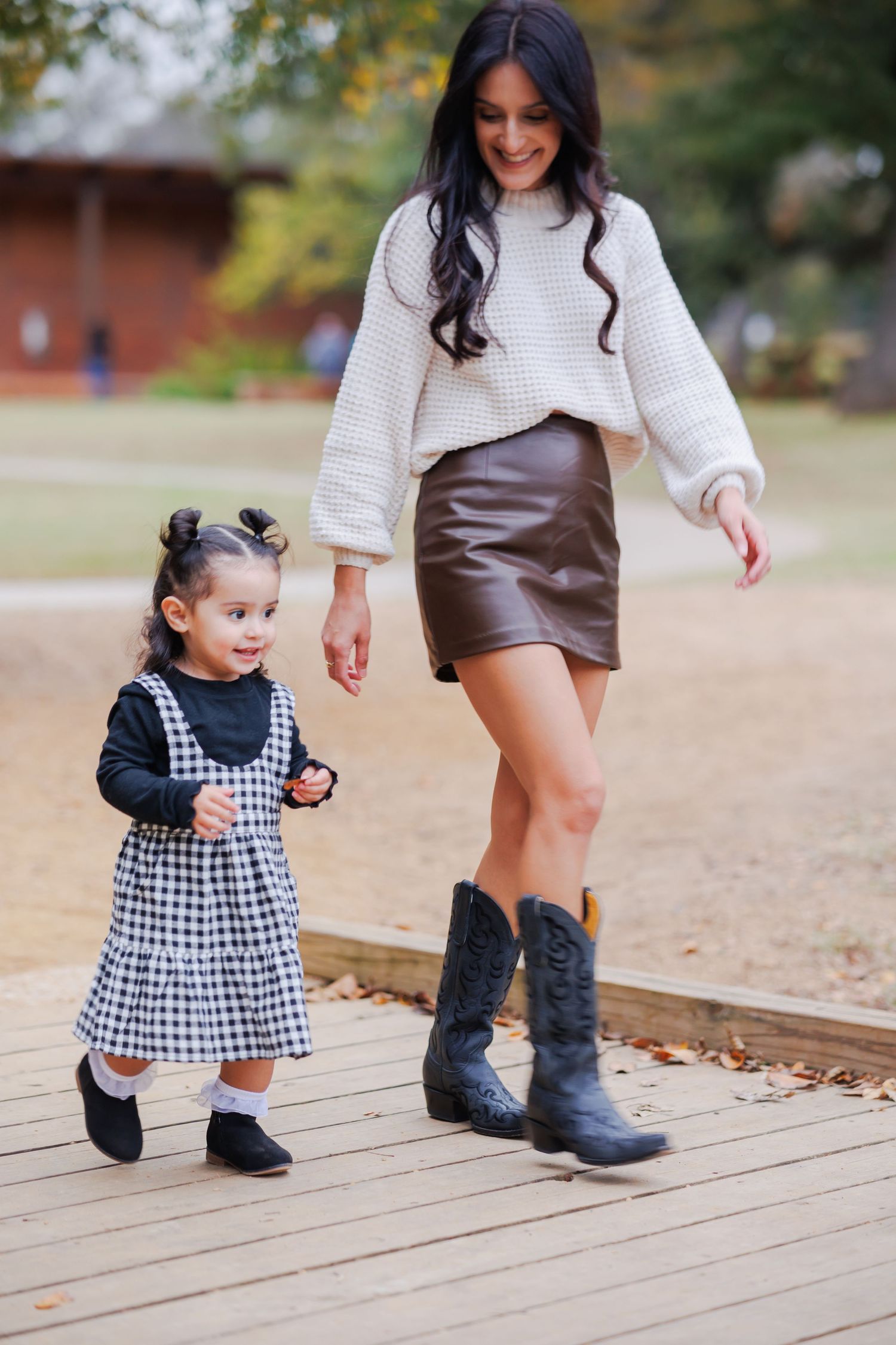 Woman in white sweater and leather skirt walks with small girl in gingham dress on wooden boardwalk during autumn.