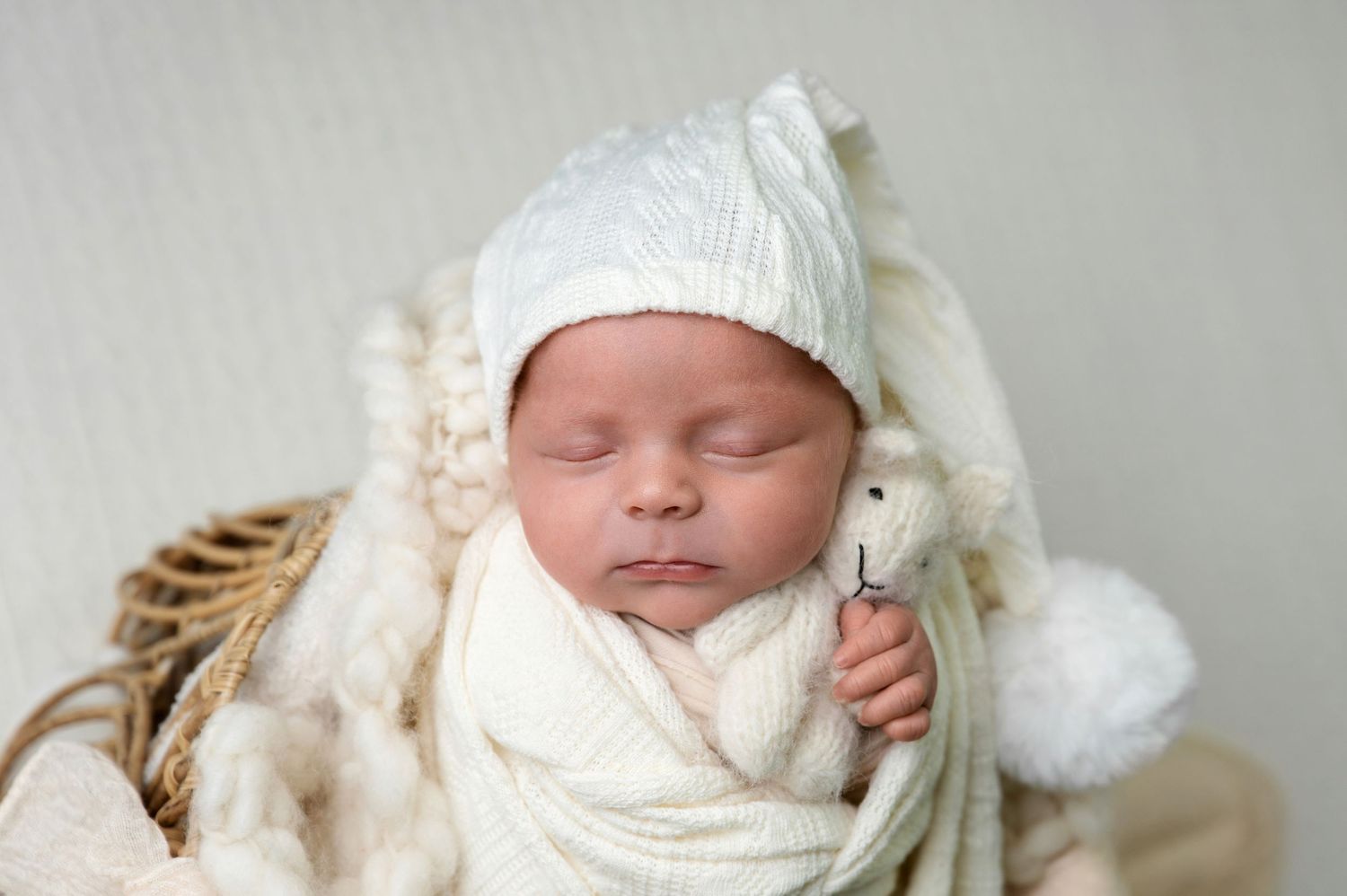 Peaceful sleeping newborn wrapped in white swaddle and knit cap rests in woven basket.