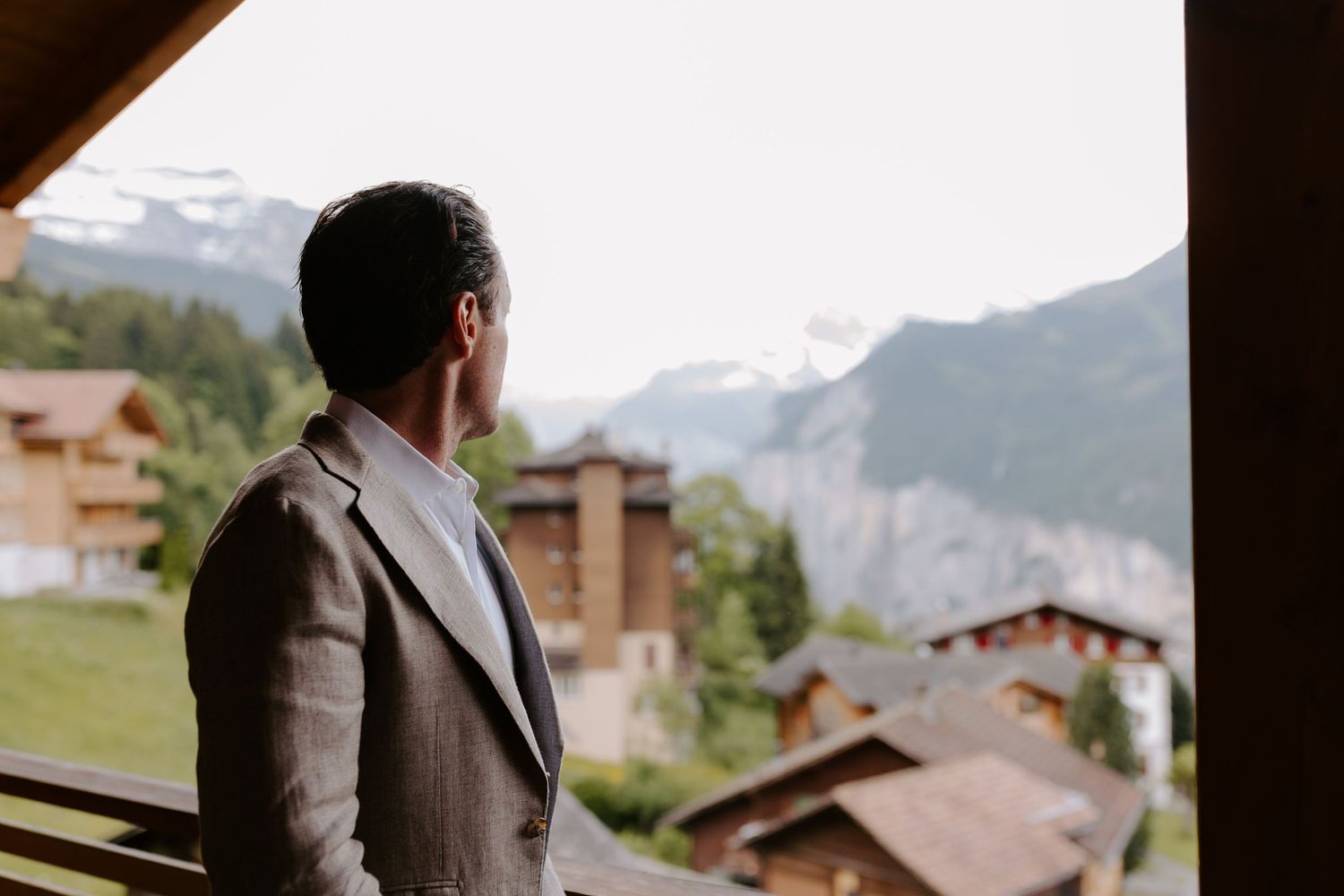 A suited figure gazes at mountain views from a chalet balcony in Switzerland.
