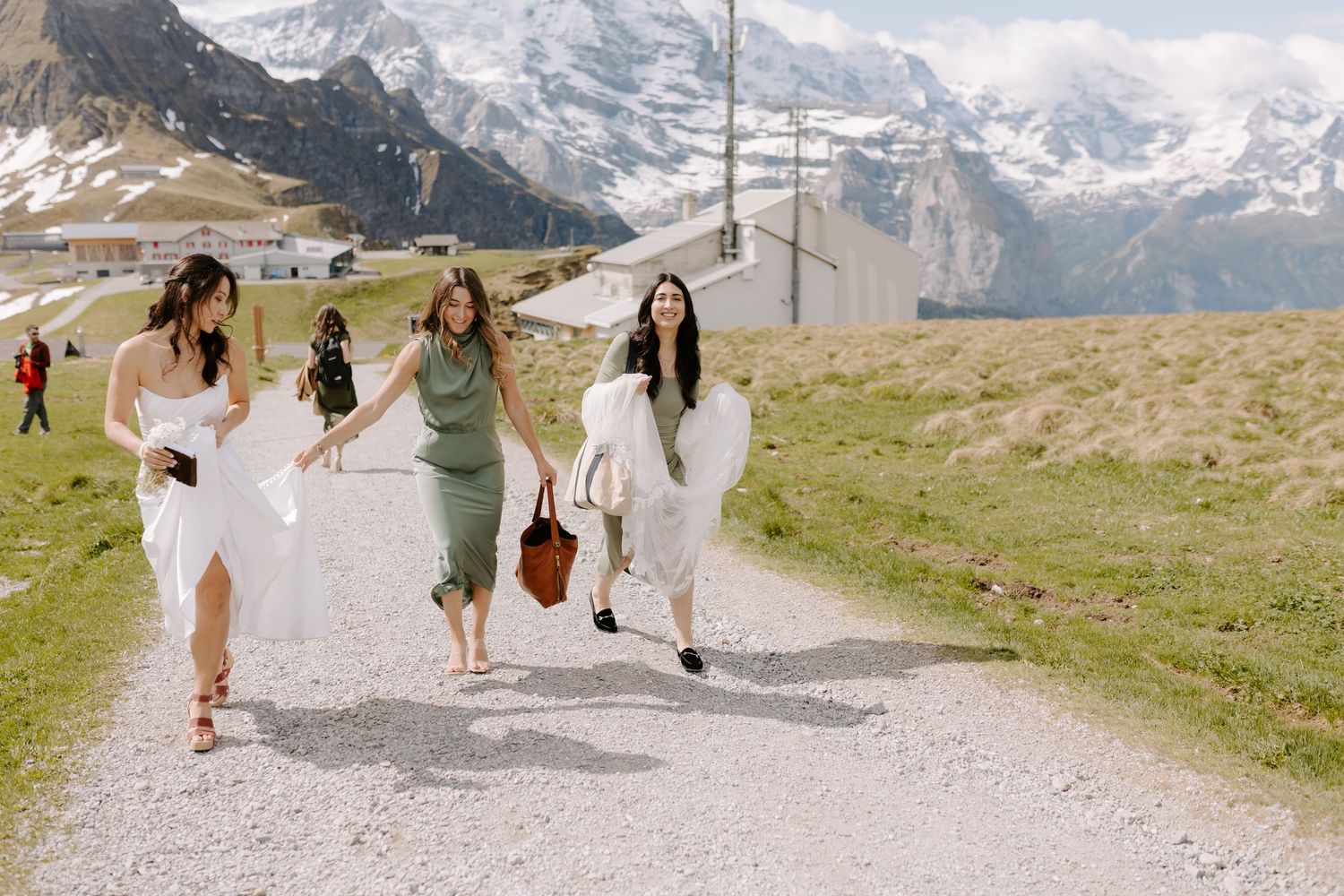 A group runs joyfully along a mountain path with snow-capped Alps in the background.
