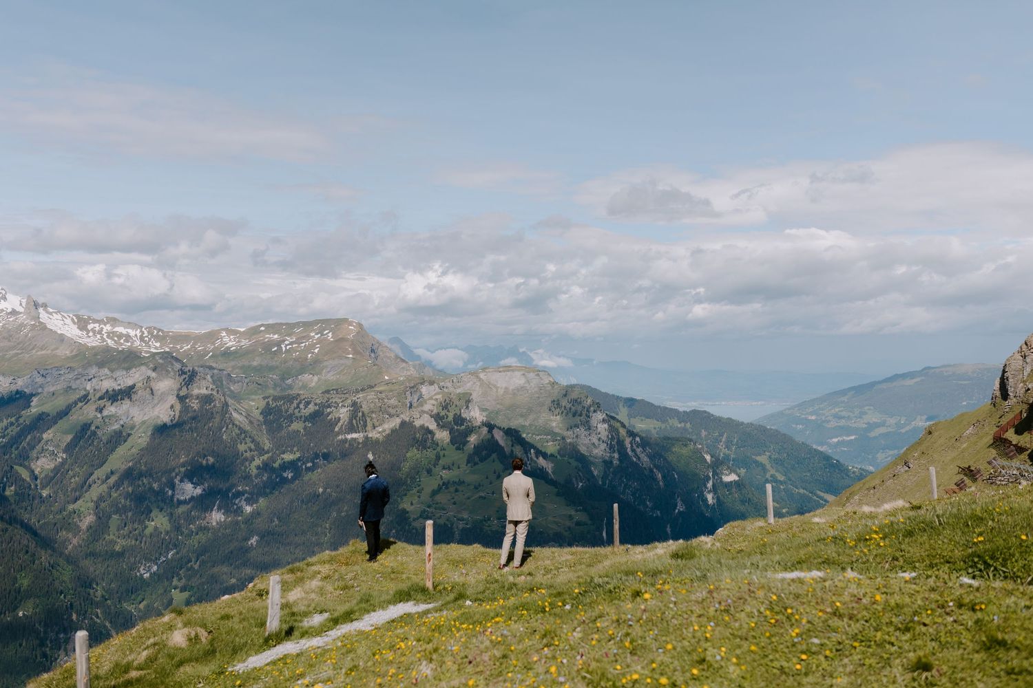 Two silhouettes stand on a grassy mountaintop overlooking a dramatic Alpine landscape.