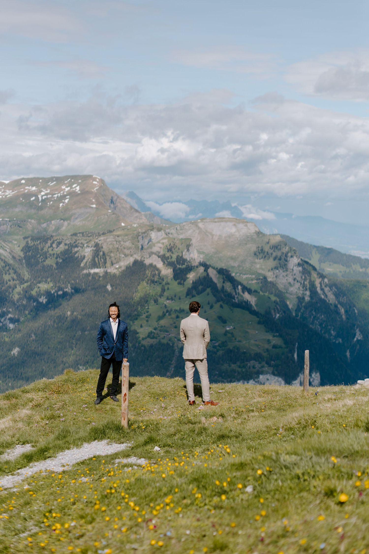 Two people stand on a mountain summit meadow with wildflowers and dramatic alpine peaks in the distance.