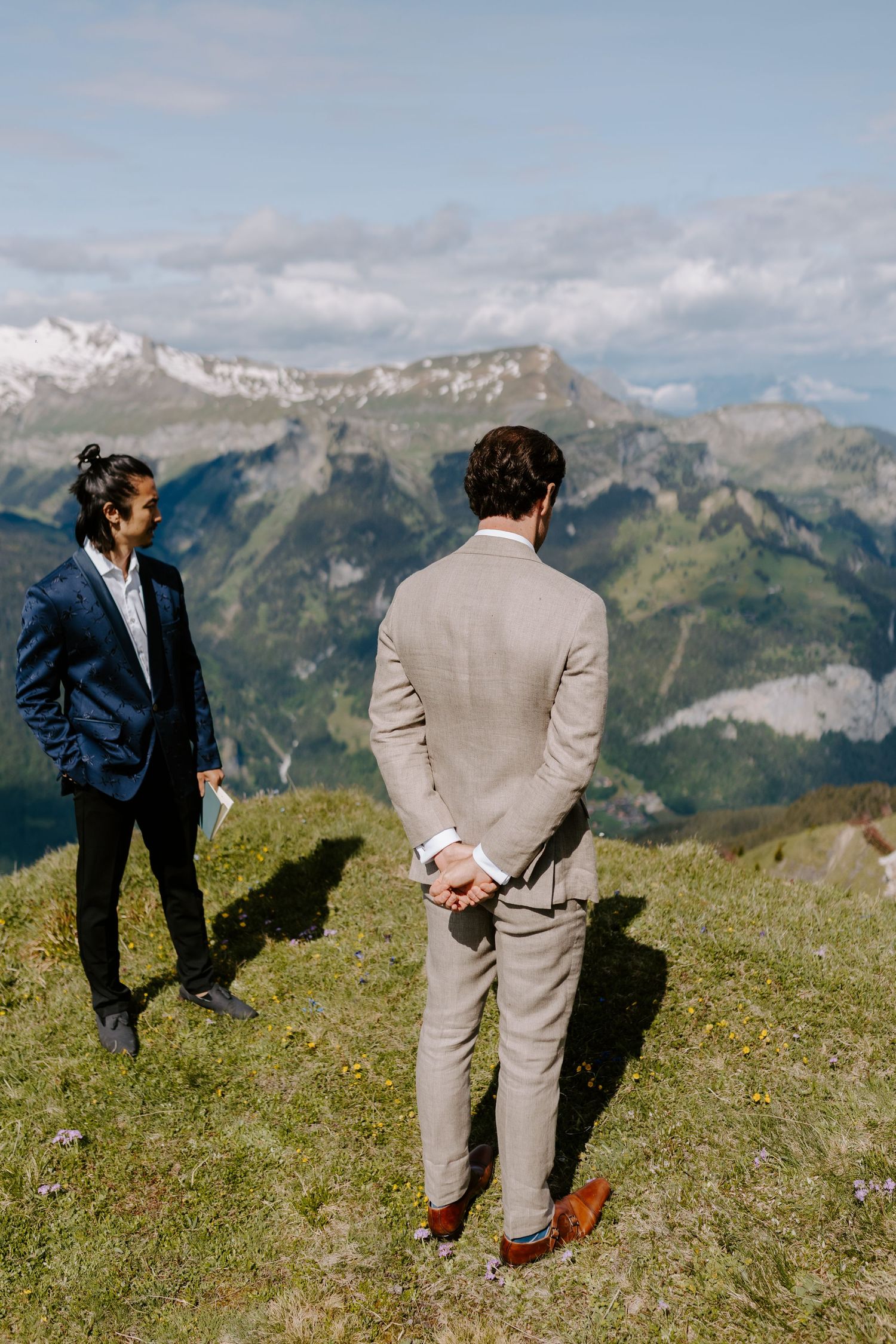 People gather on a grassy alpine meadow surrounded by rugged mountain peaks on a sunny day.