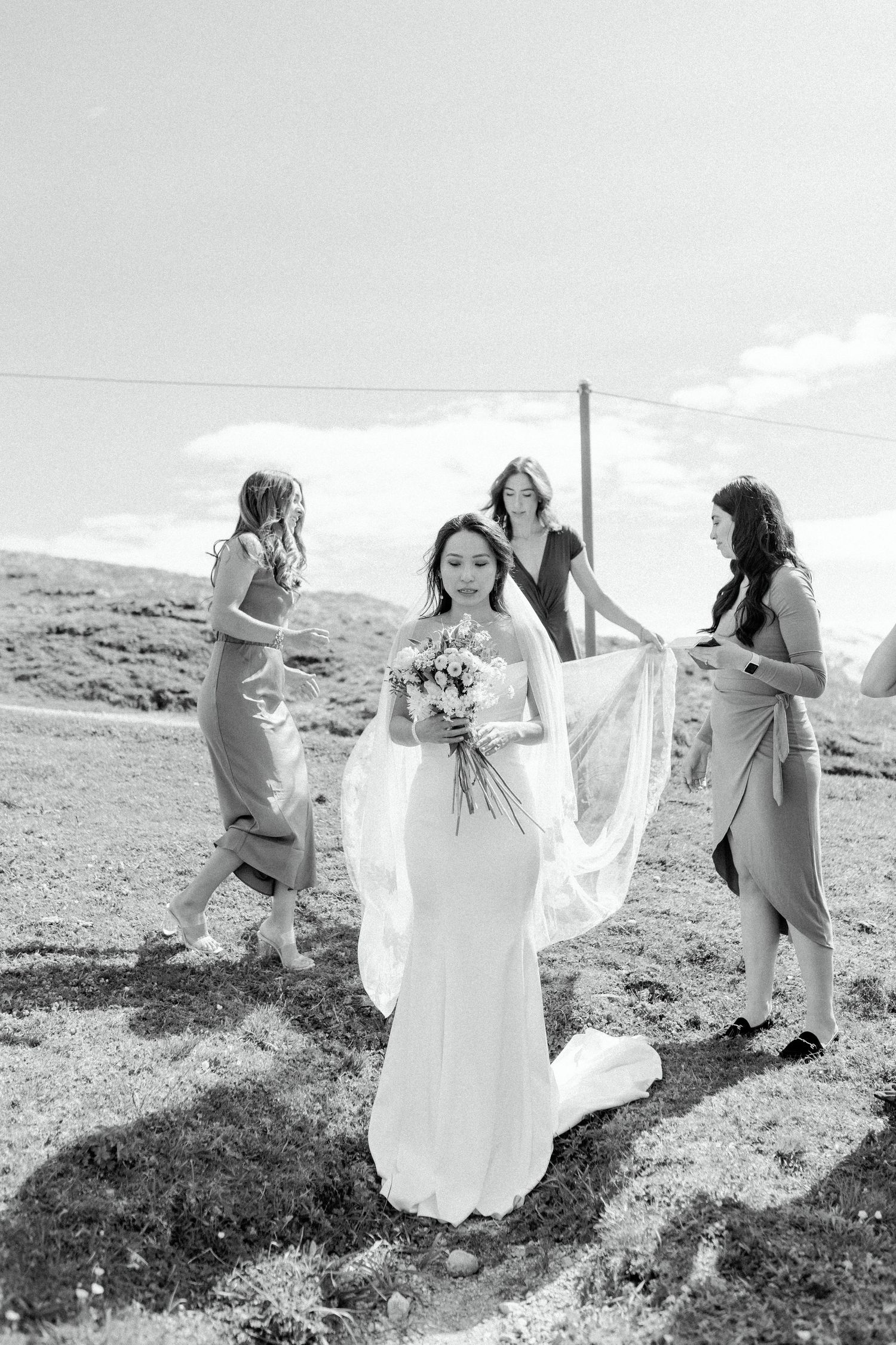 Bride in flowing white dress surrounded by bridesmaids helping with her gown on a grassy mountain hillside.