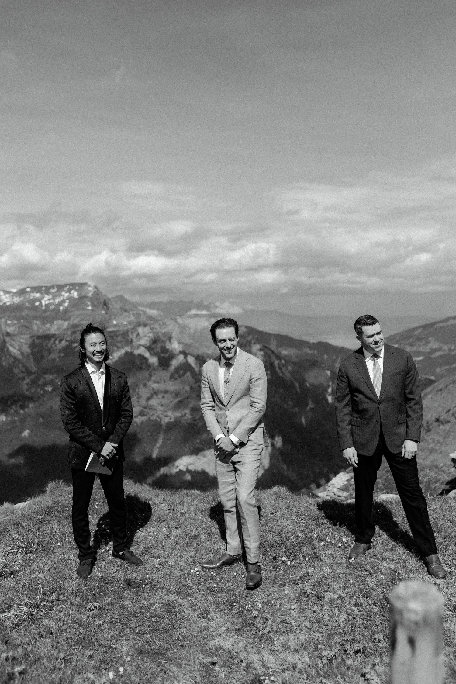 Three groomsmen in suits posing casually against dramatic mountain backdrop in black and white photography.