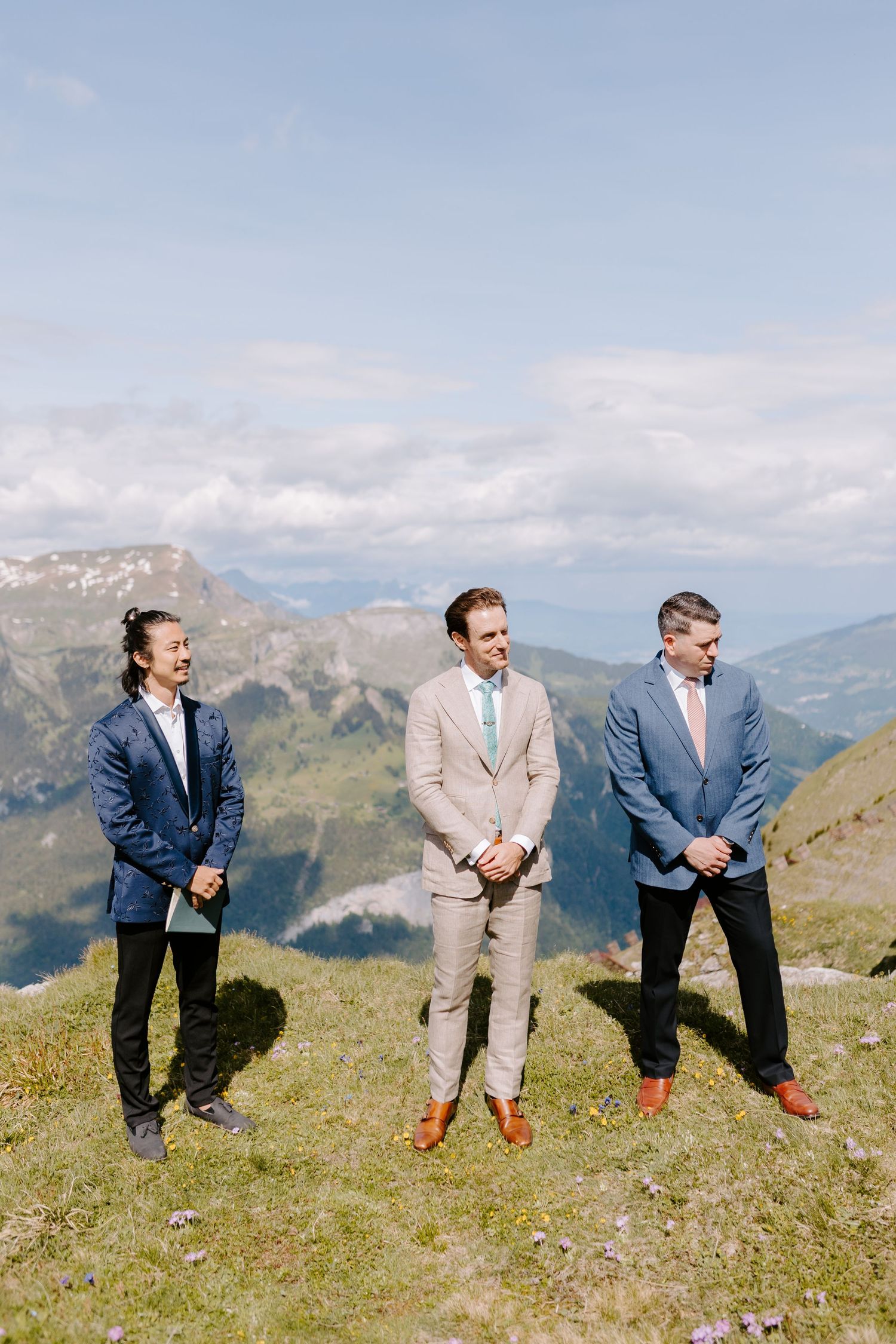 Three groomsmen stand on a grassy mountain summit against a backdrop of majestic Alpine peaks.
