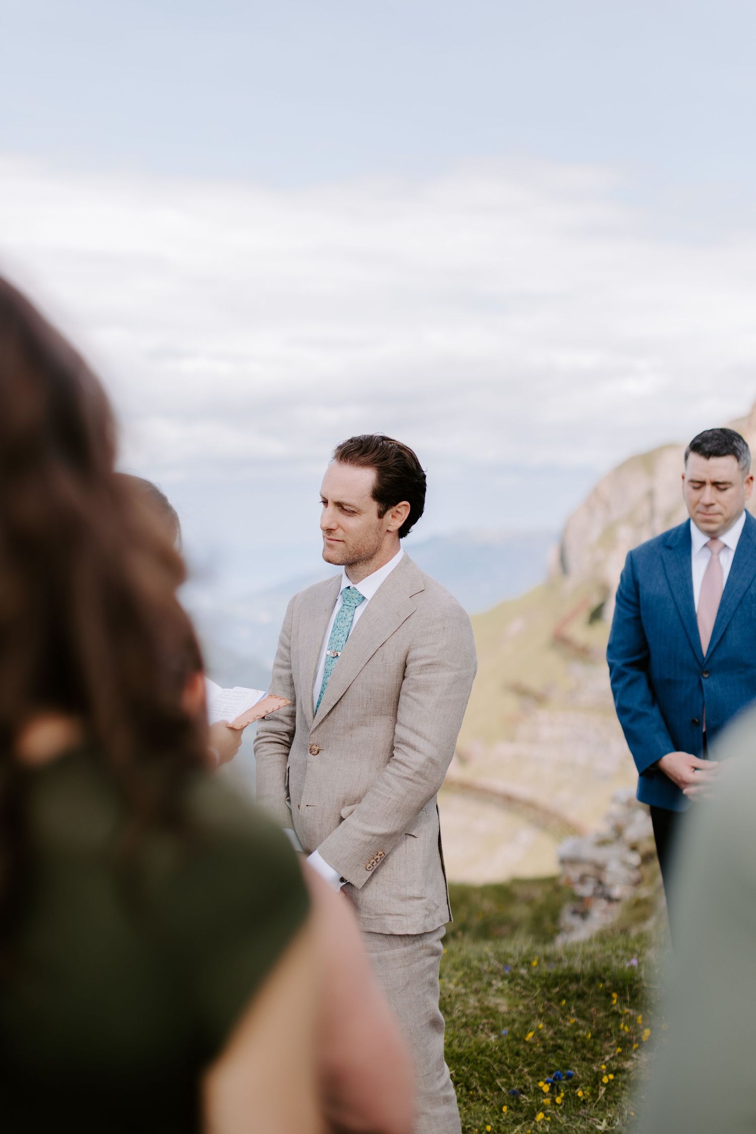 A wedding ceremony takes place on a scenic cliff overlooking mountains, with suits in tan and navy blue.