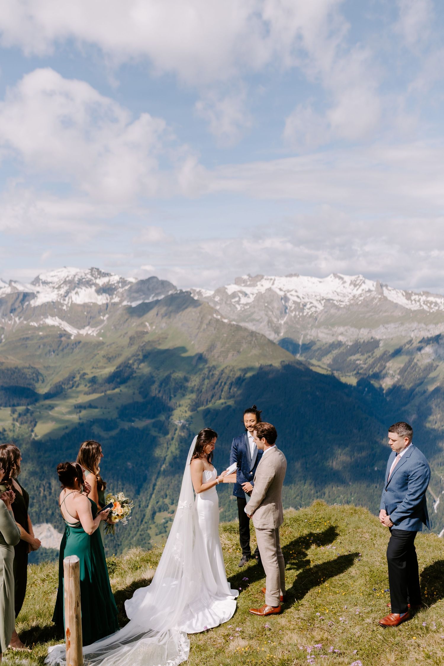 A small wedding ceremony takes place on a mountaintop with snow-capped peaks and blue skies in the background.