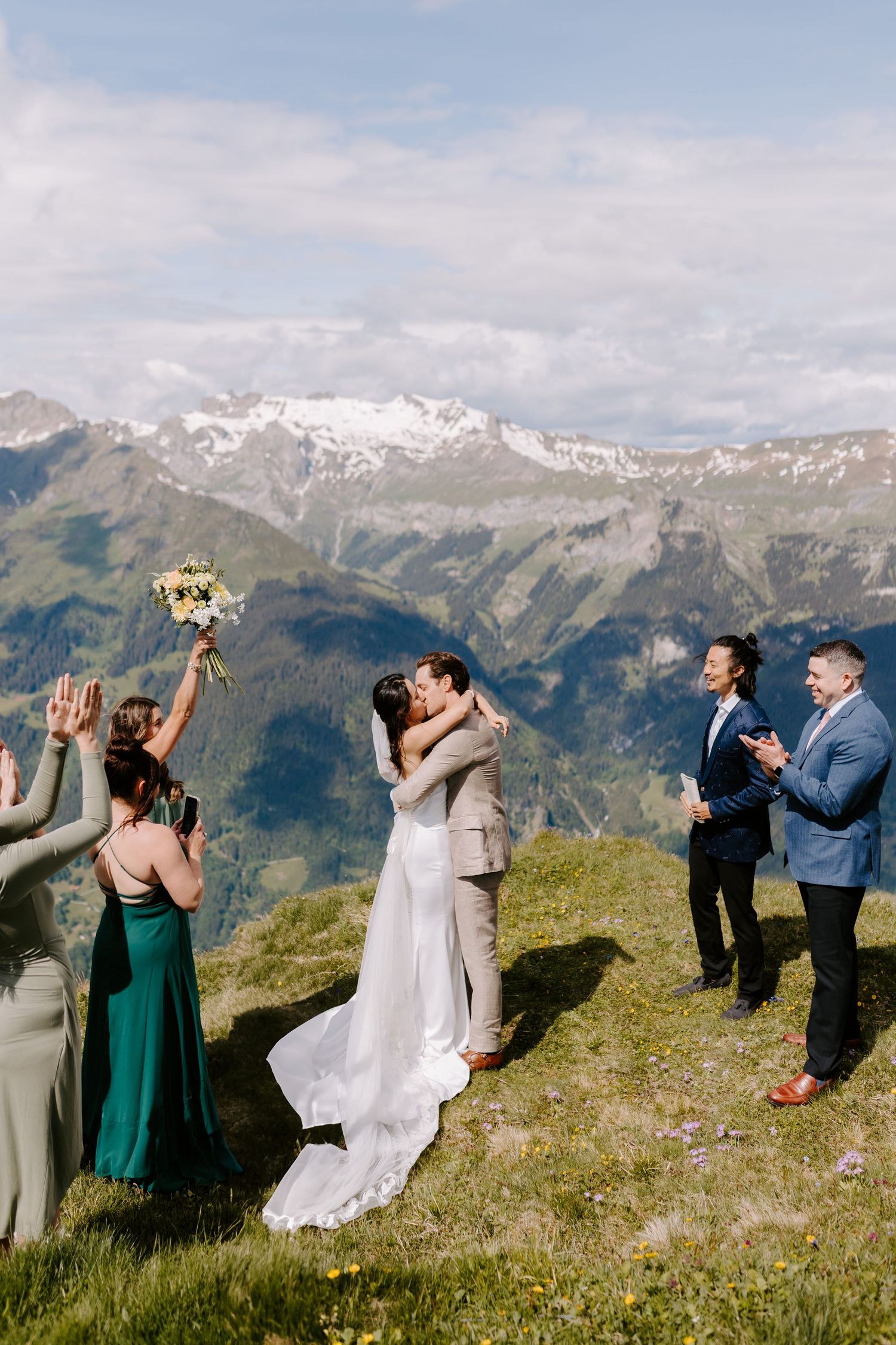 A mountain wedding ceremony takes place on a scenic alpine meadow with snow-capped peaks in the background.