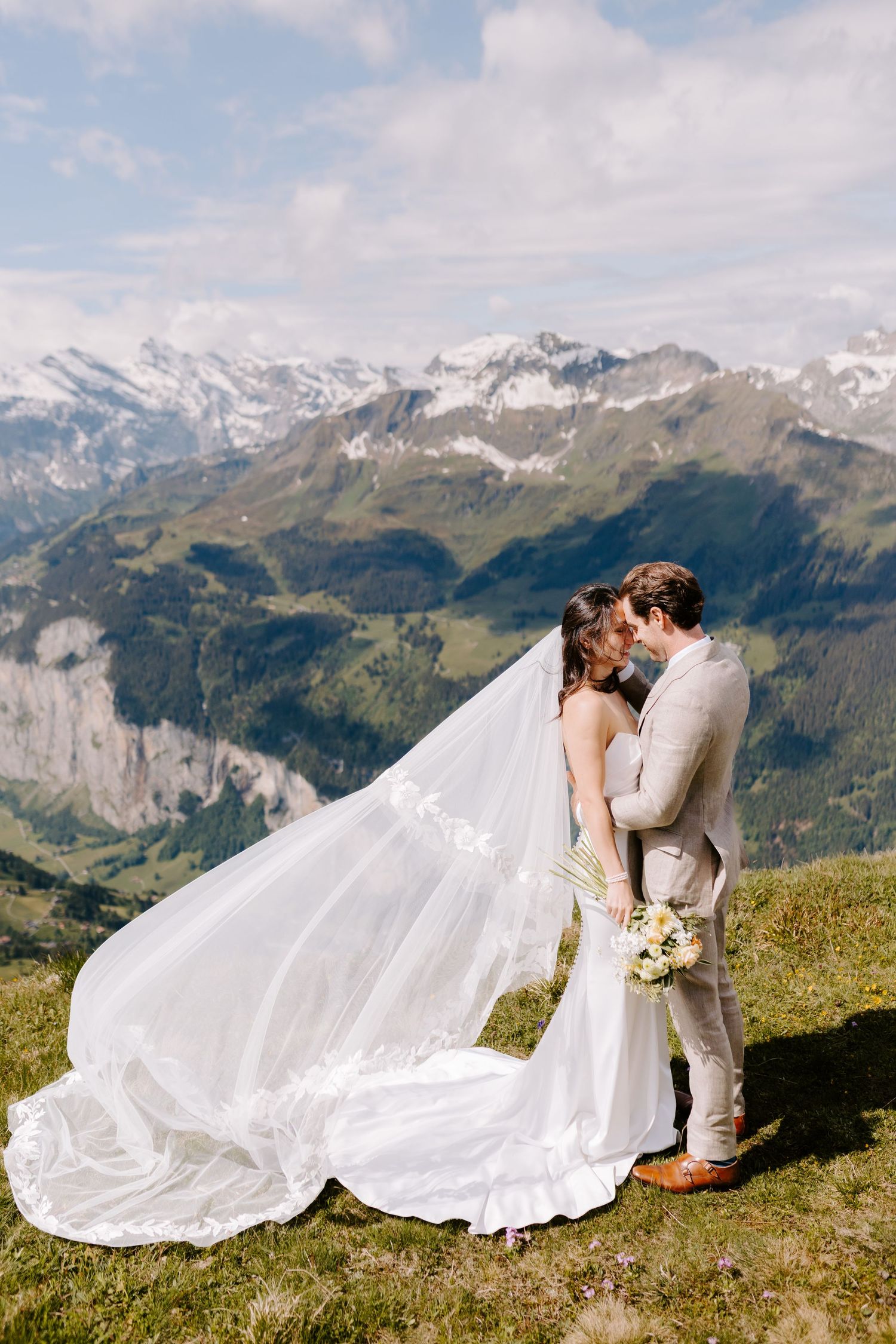 Newlyweds share romantic kiss on mountaintop with flowing wedding dress and stunning alpine vista.